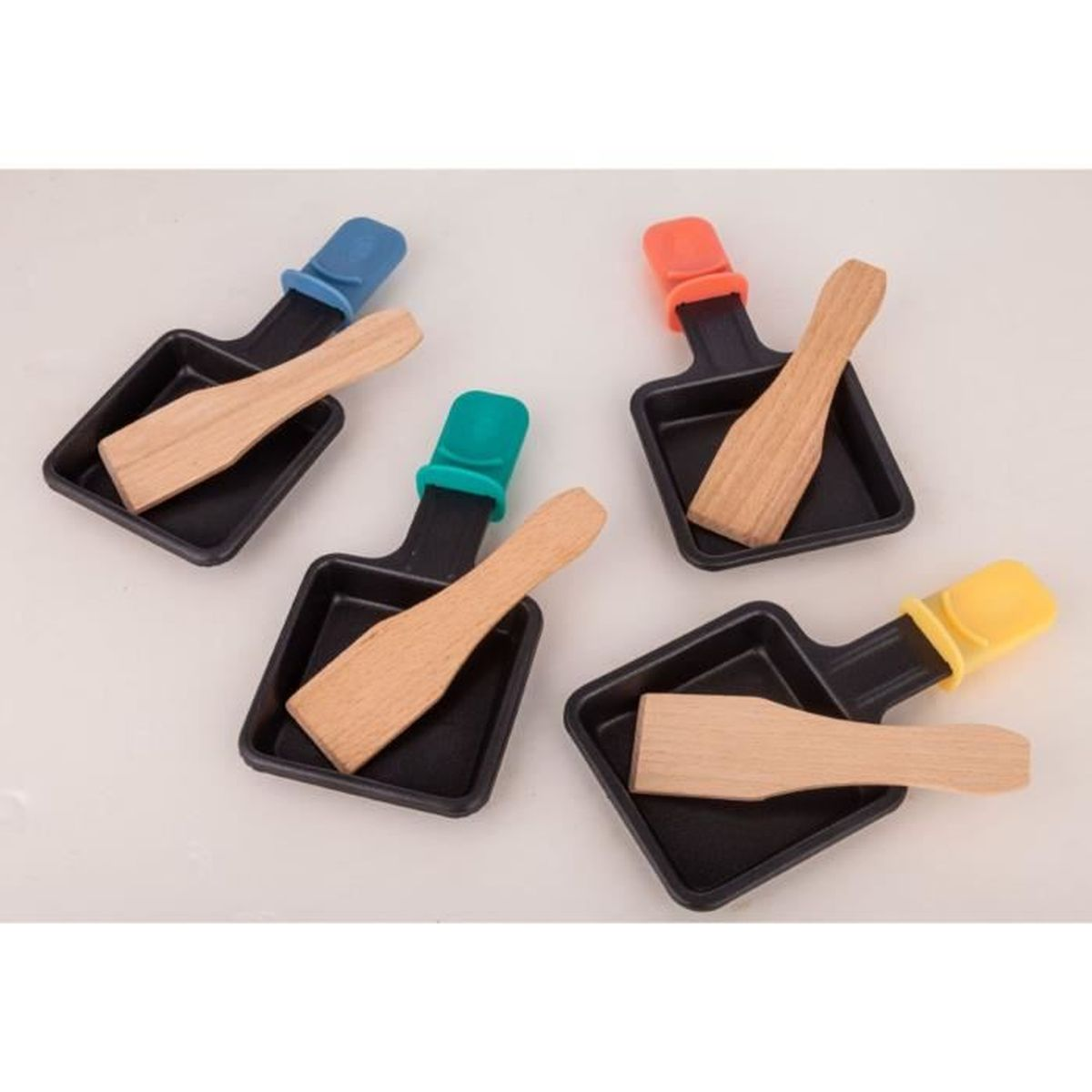 Fünf Raclette-Grillpfannen mit Holzspateln. Jede Pfanne hat einen farbigen Griff: blau, grün, orange, gelb. Auf einer weißen Oberfläche.