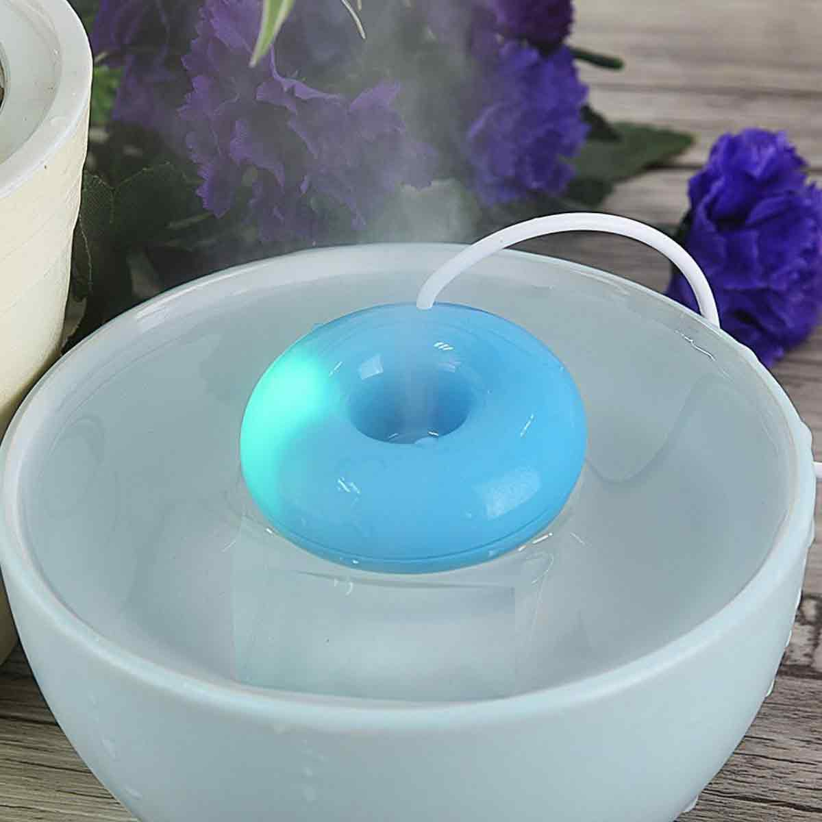 Ein blauer, donutformiger Luftbefeuchter schwimmt in einer Wasserschüssel. Lila Blumen und Dampf im Hintergrund.