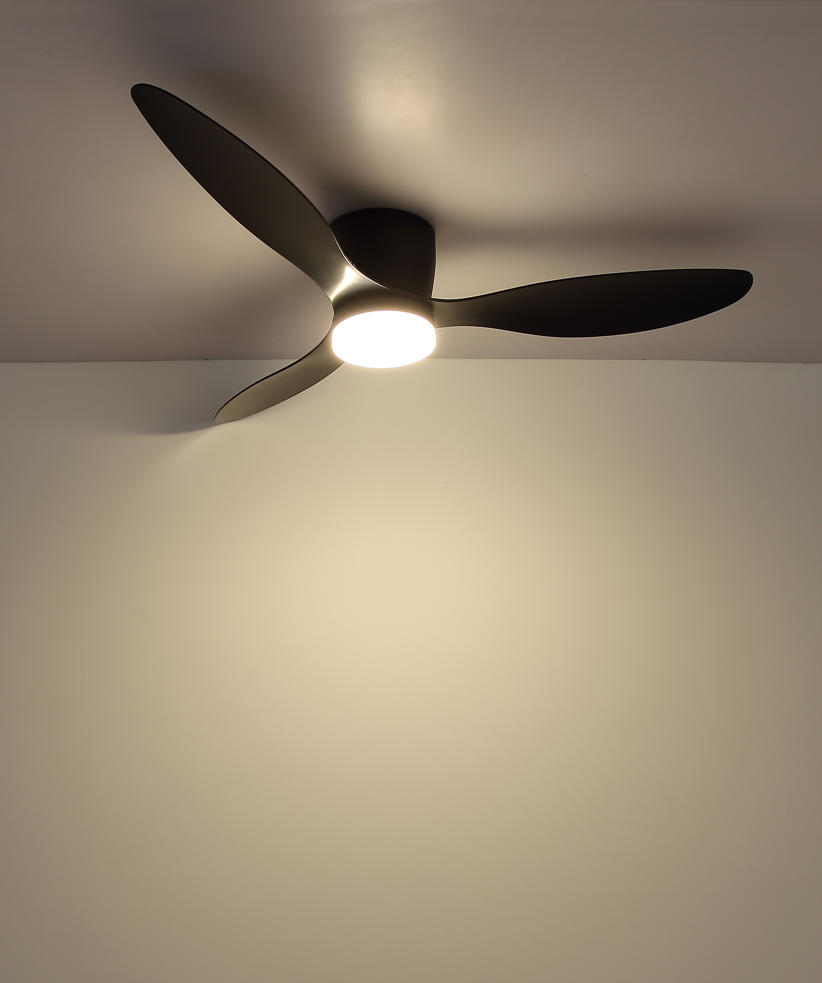 Ein Deckenventilator mit drei schwarzen Blättern und einem zentralen Licht ist an einer weißen Decke montiert. Das Licht ist an.