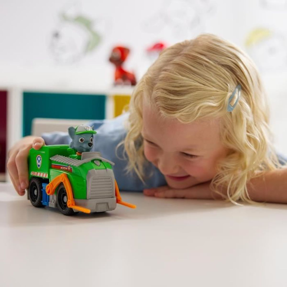 Ein Mädchen lächelt einen grünen Spielzeug-LKW mit einer Zeichentrickfigur an. Der LKW steht auf einer weißen Oberfläche. Der Hintergrund hat Spielzeug und bunte Formen.