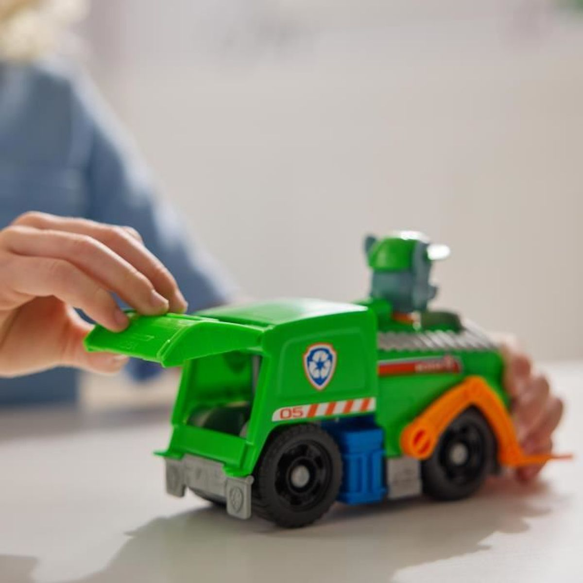 Hände spielen mit einem grünen Spielzeug-Müllwagen. Der Truck hat schwarze Räder, orangefarbene Arme und blaue und weiße Details.