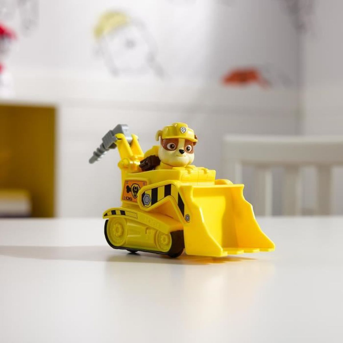 Ein gelber Spielzeugbagger mit einer Cartoon-Hundefigur am Steuer, auf einem weißen Tisch, mit verschwommenem Hintergrund.