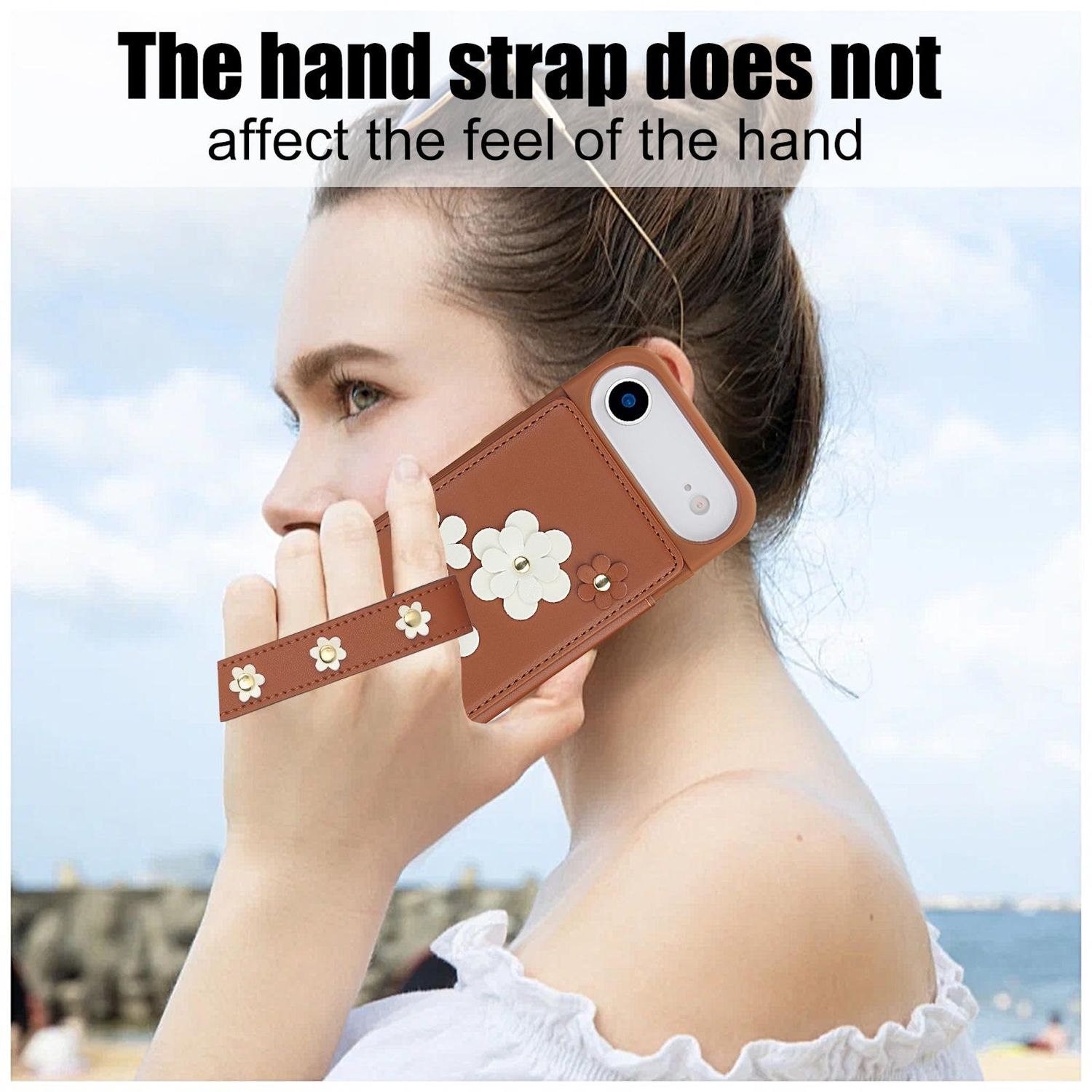 Una mujer sostiene una funda de teléfono marrón con una correa de mano. Flores blancas adornan la funda y la correa. El fondo es un cielo azul.