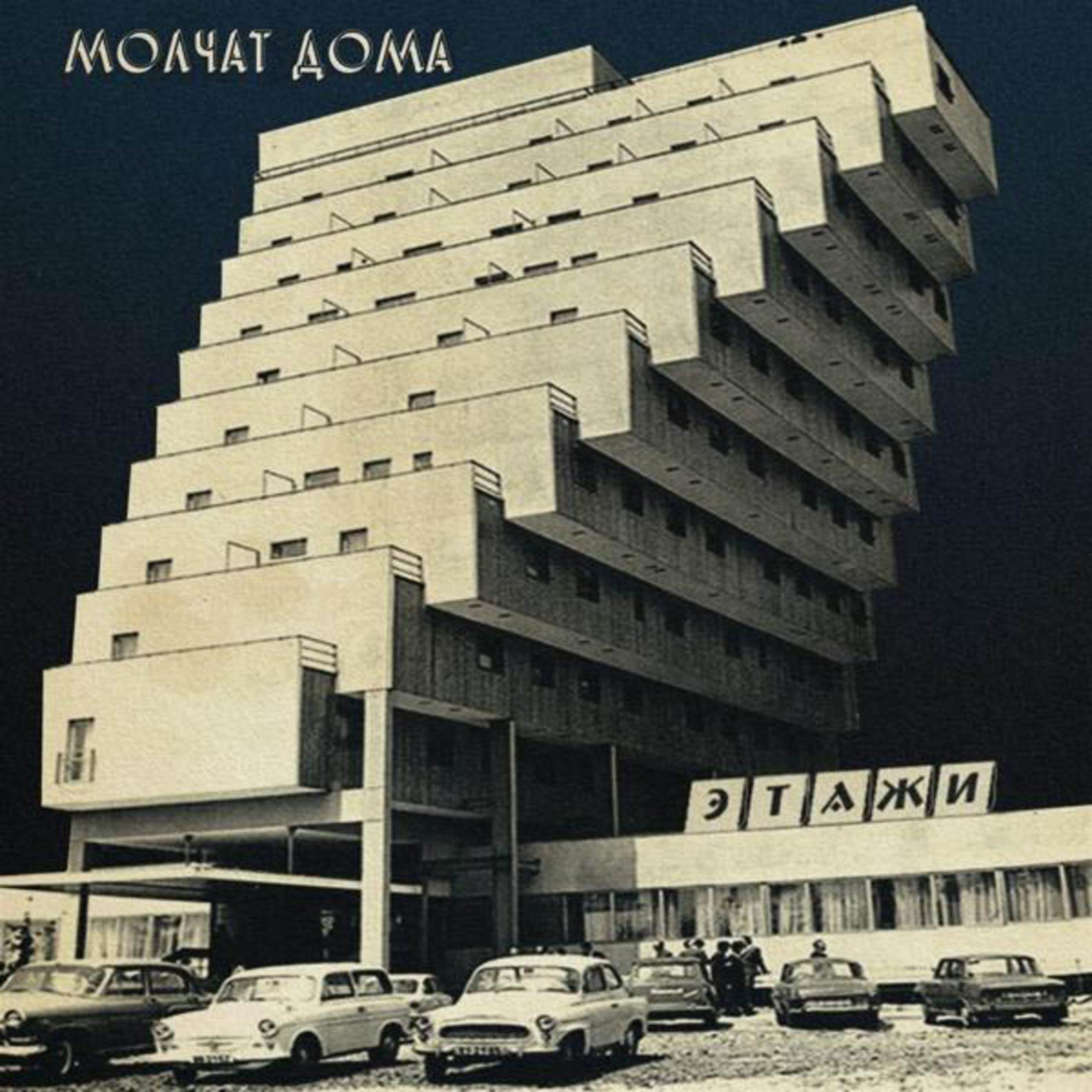 Foto d'epoca di un edificio modernista con facciata a gradini. Auto parcheggiate davanti. Testo in russo in alto e in basso.