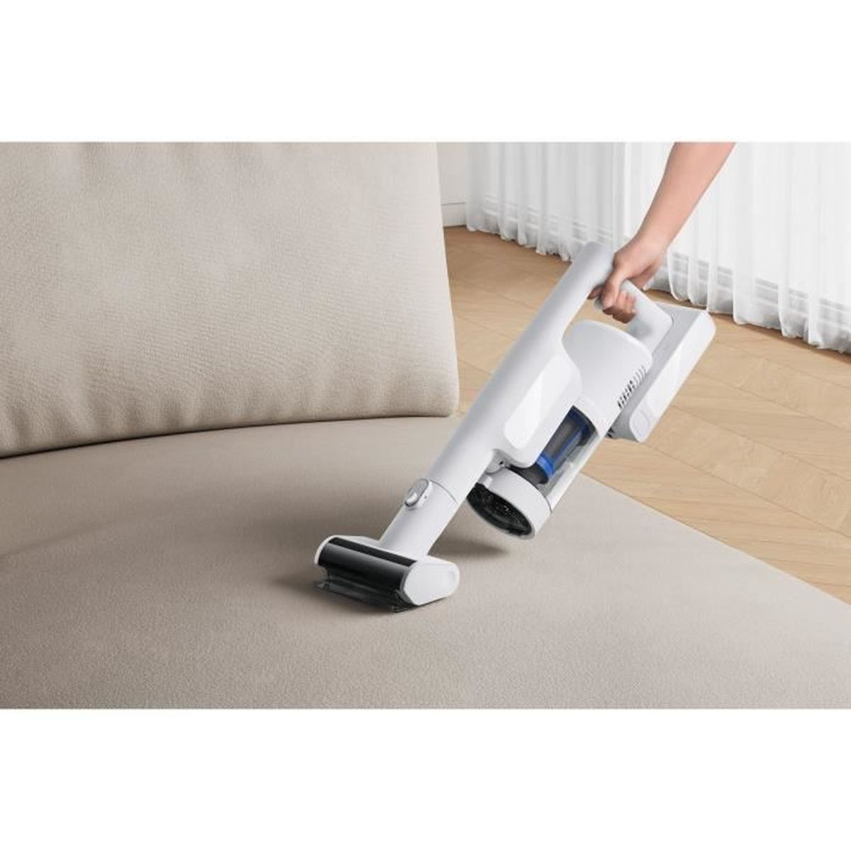 Une main tient un aspirateur sans fil blanc, nettoyant un canapé beige. L'arrière-plan montre un plancher en bois et un rideau blanc.