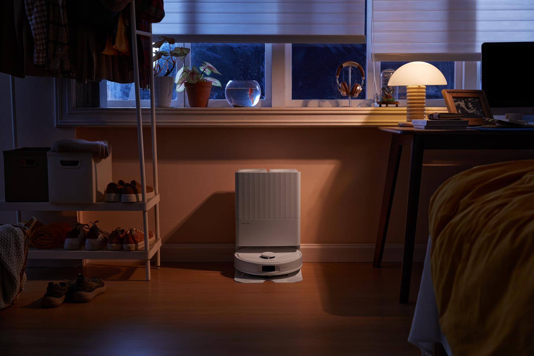 A room with a robotic vacuum cleaner, a window, a table with a lamp, and a bed covered with a yellow blanket.