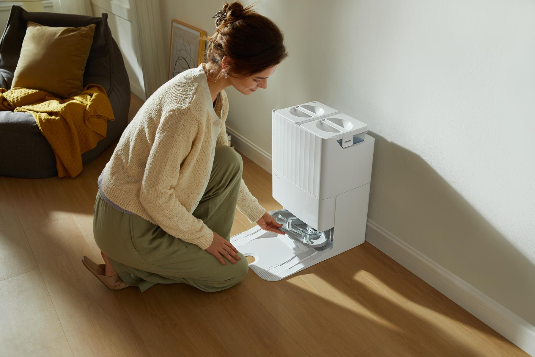 Woman placing a white cleaning device component on a wooden floor. A beanbag and a yellow blanket are in the background.