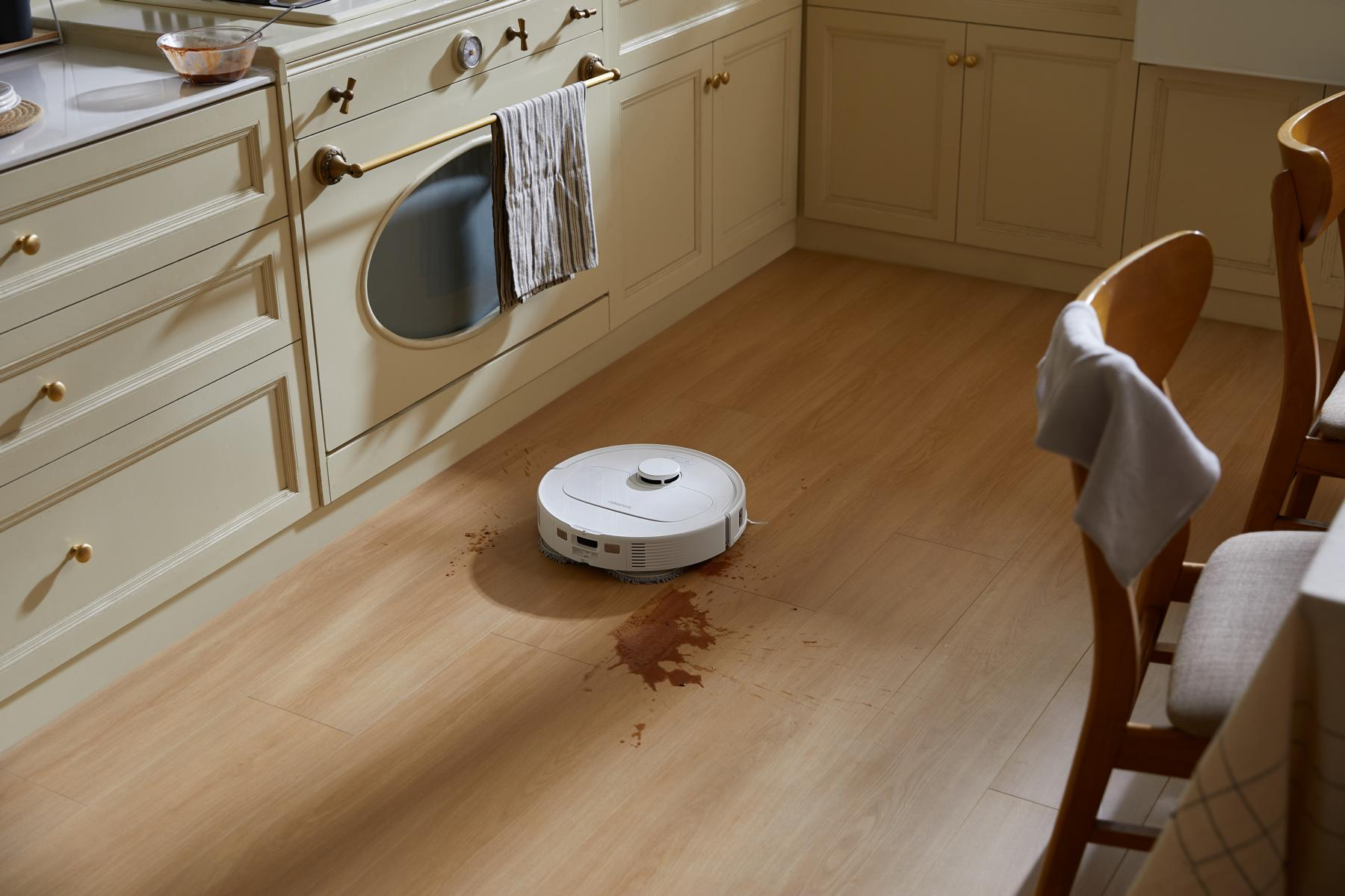 A robot vacuum cleans a kitchen floor with a spill. Cabinets and chairs are in the background.