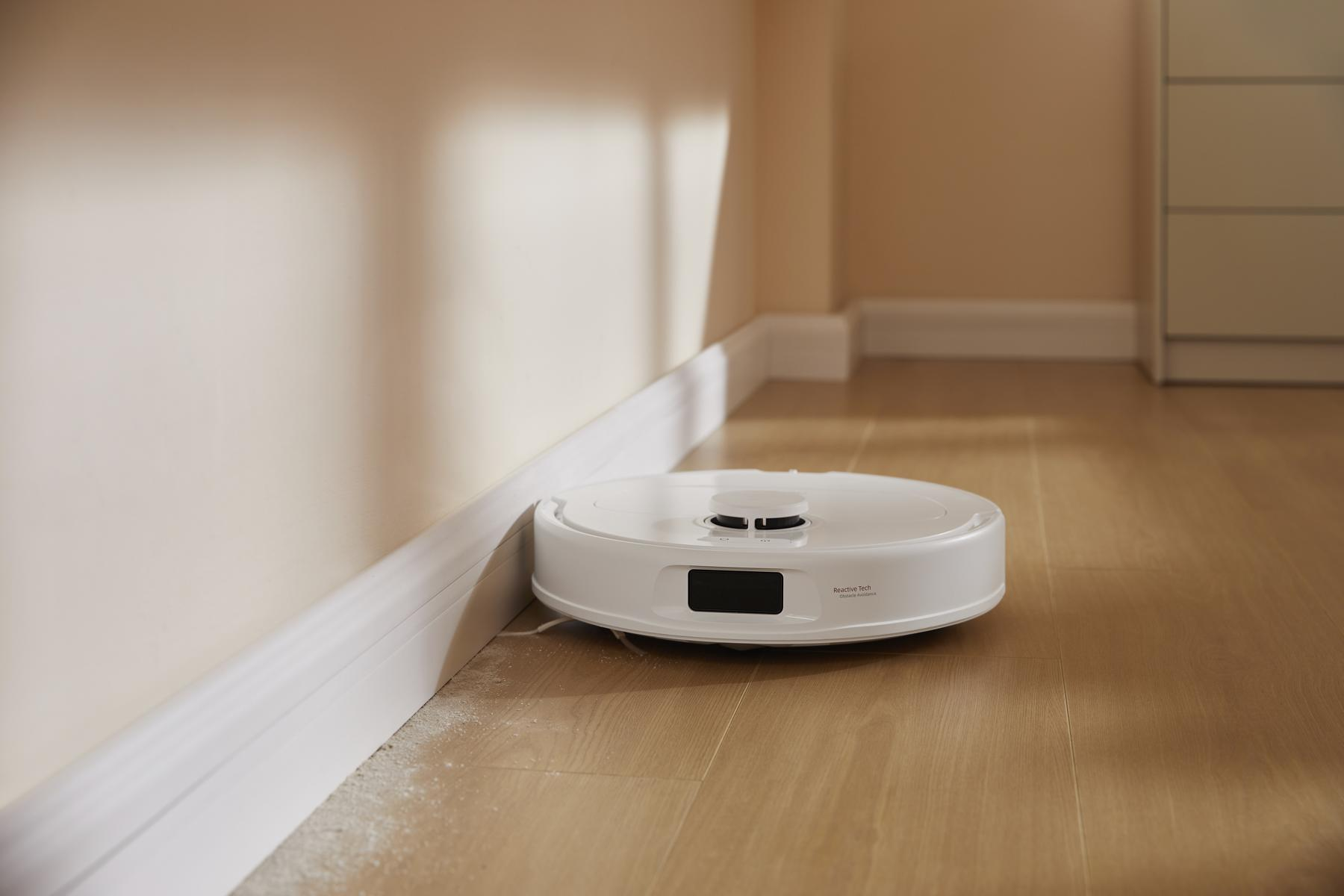 A white robot vacuum cleaner on a wooden floor near a wall. The wall is beige, and the floor is wood-colored.