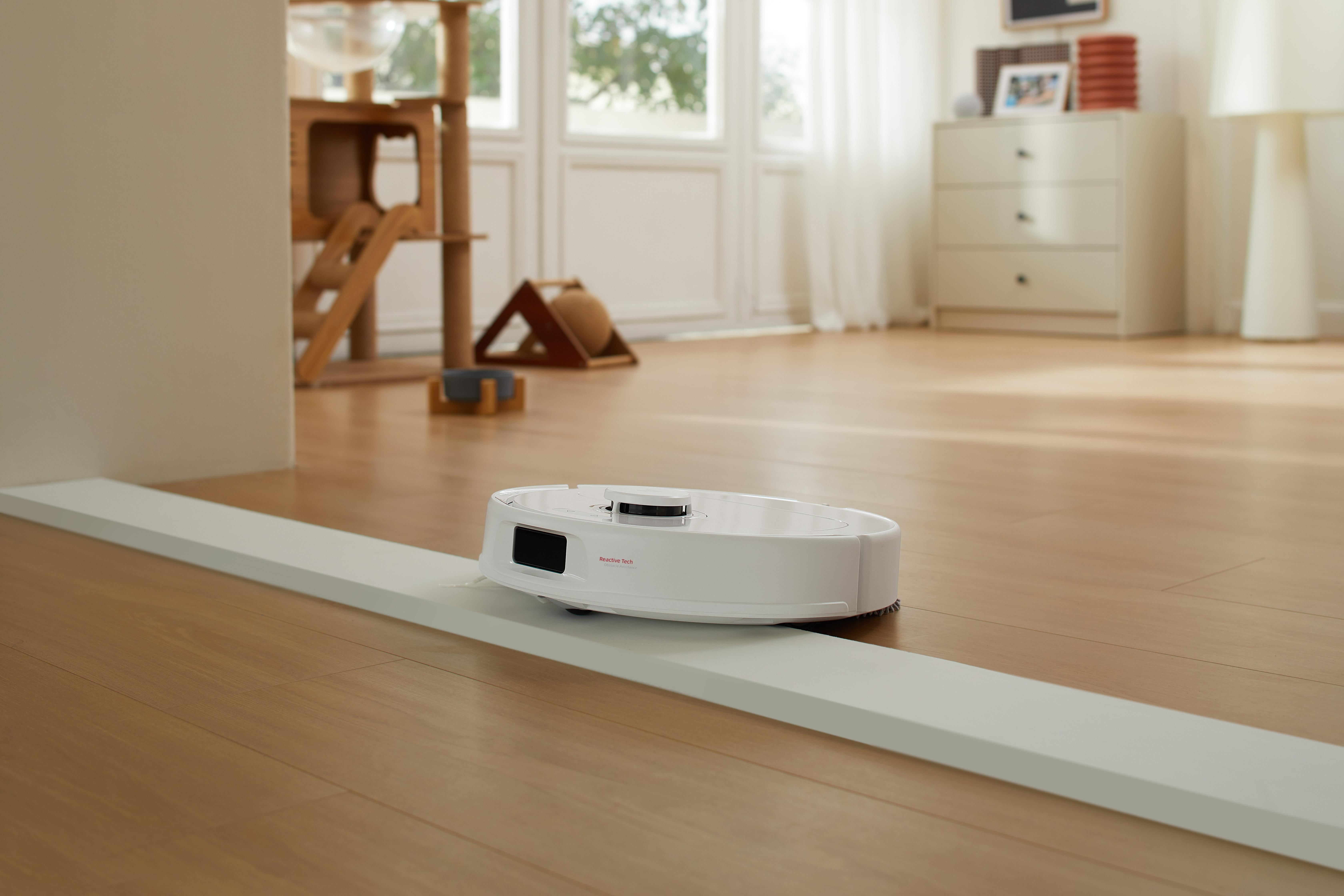 A white robot vacuum cleaner on a wooden floor, near a white barrier. A cat tree and furniture are in the background.