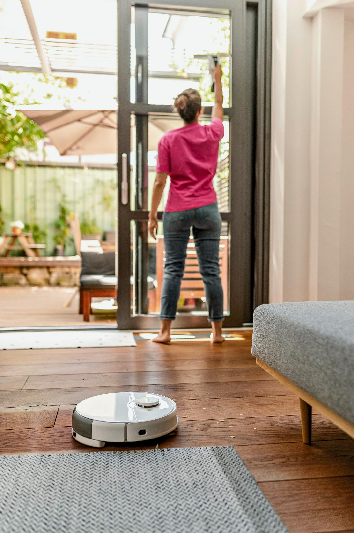 Kobieta czyści szklane drzwi. Robot odkurzający stoi na drewnianej podłodze. Widoczna jest również sofa i miejsca do siedzenia na zewnątrz.