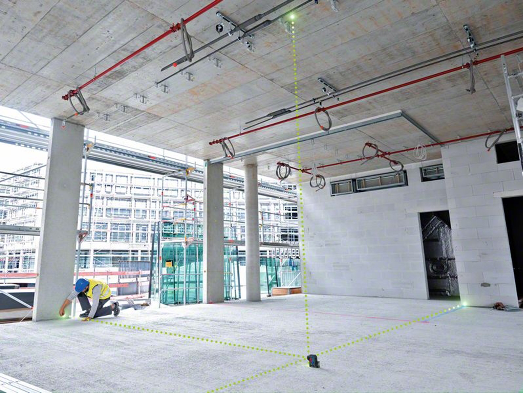 Bir inşaat işçisi diz çökmüş, lazer seviyesi kullanıyor. Odada beton sütunlar, kırmızı bir boru sistemi ve beyaz tuğla bir duvar var.