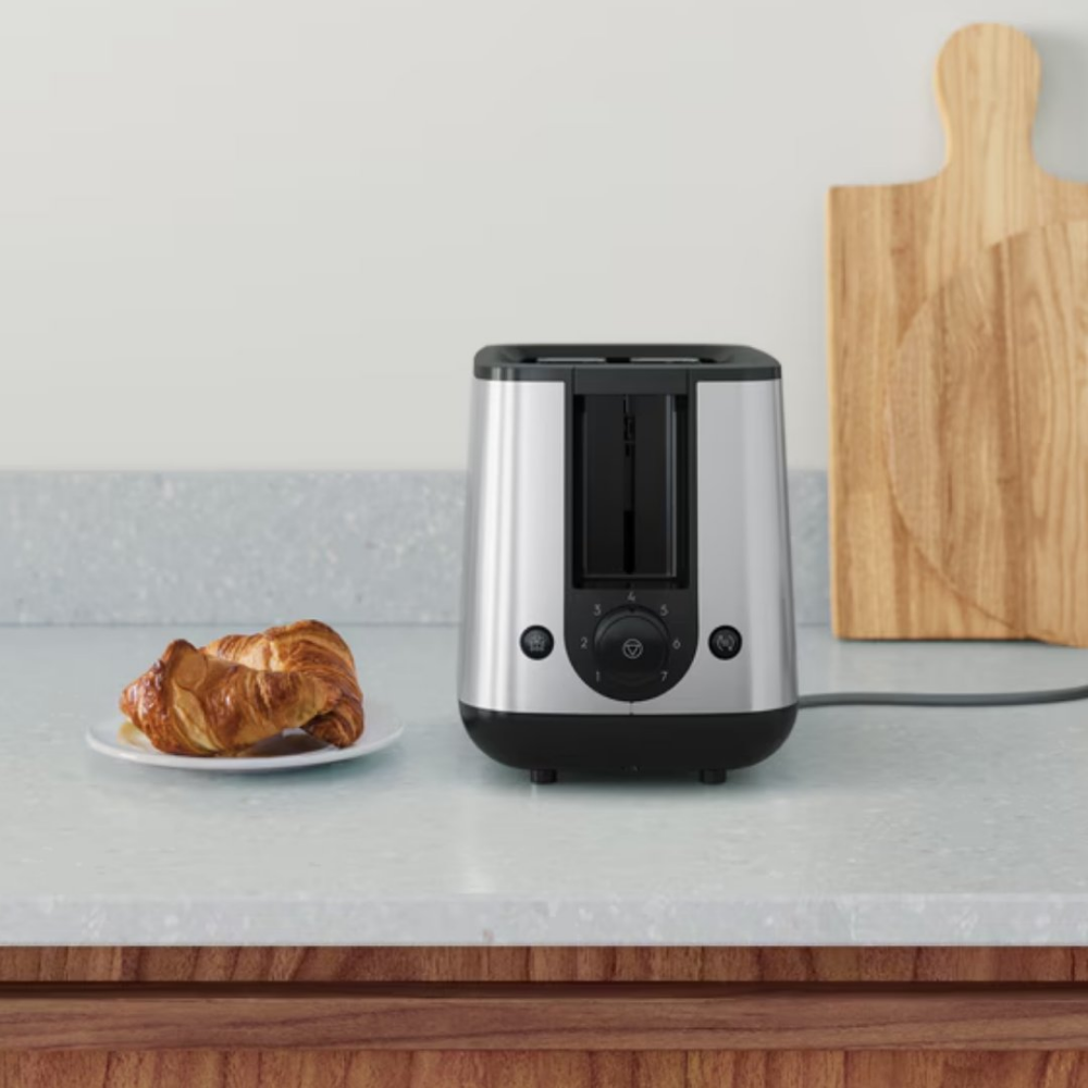 Toster ze stali nierdzewnej stoi obok talerza z rogalikiem. Drewniana deska do krojenia jest w tle.