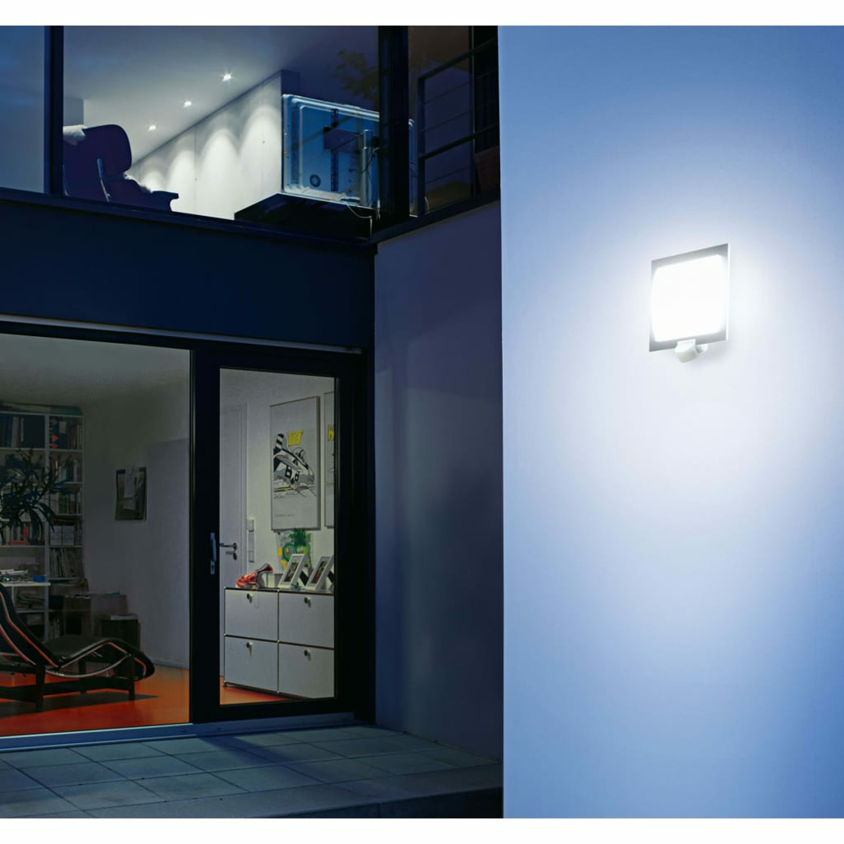 Außenansicht eines Hauses in der Dämmerung. Eine beleuchtete Wandlampe, große Fenster und Innendetails wie Möbel und Kunst sind sichtbar.