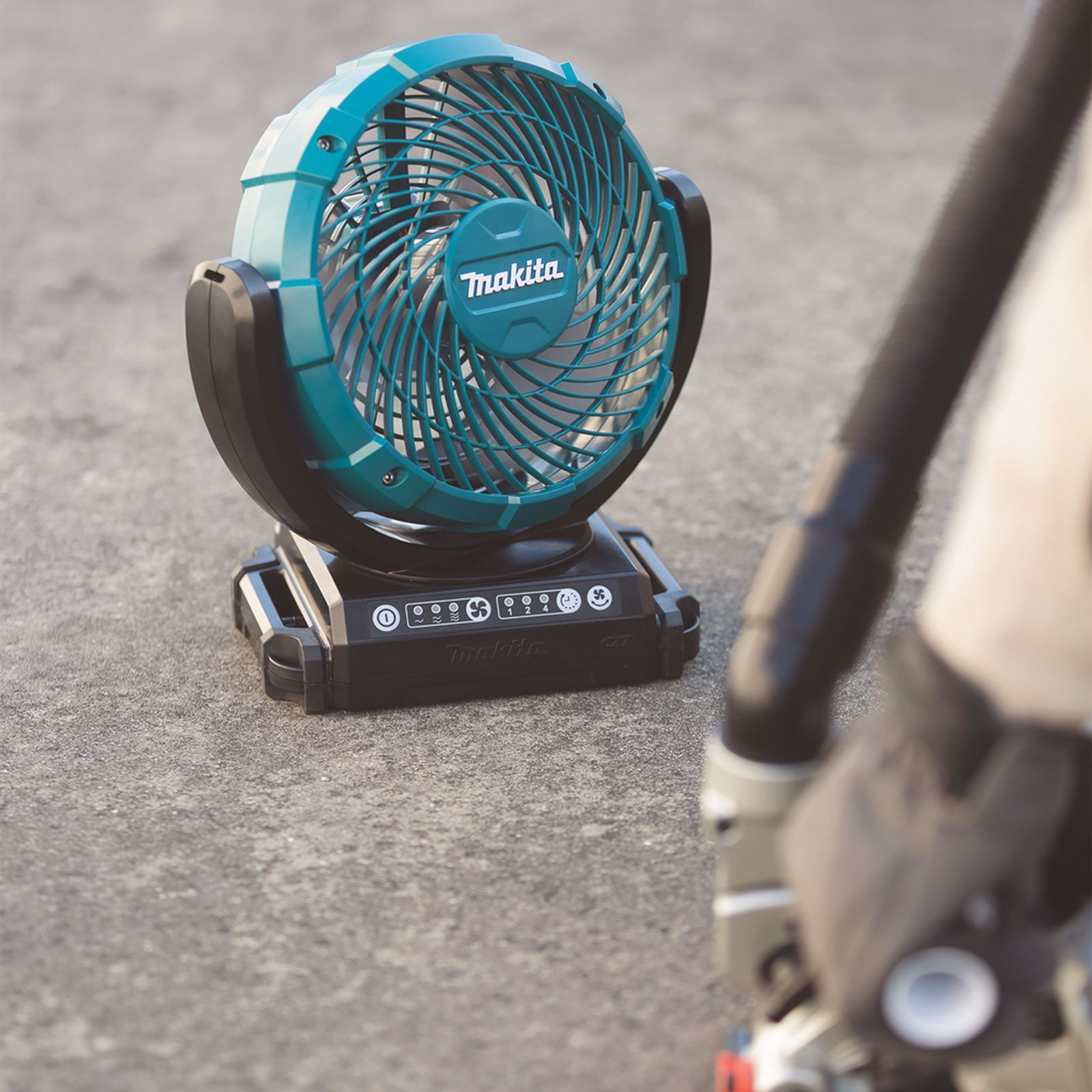 Un ventilatore Makita turchese e nero è su cemento. Una persona con un tubo nero è in primo piano sfocato.