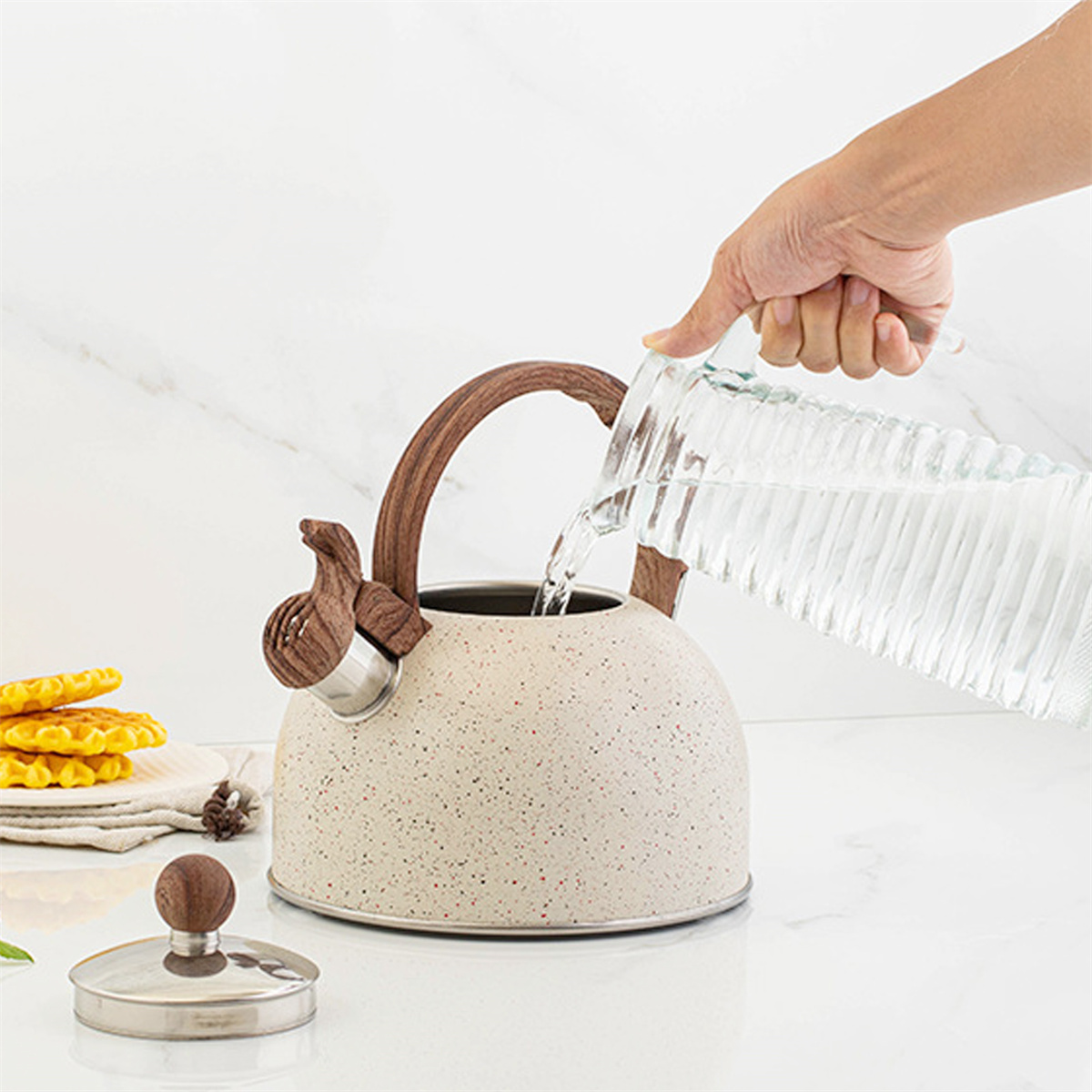Eine Hand gießt Wasser aus einem Glaskrug in einen gesprenkelten Wasserkocher. Waffeln und der Kesseldeckel liegen auf der Theke.