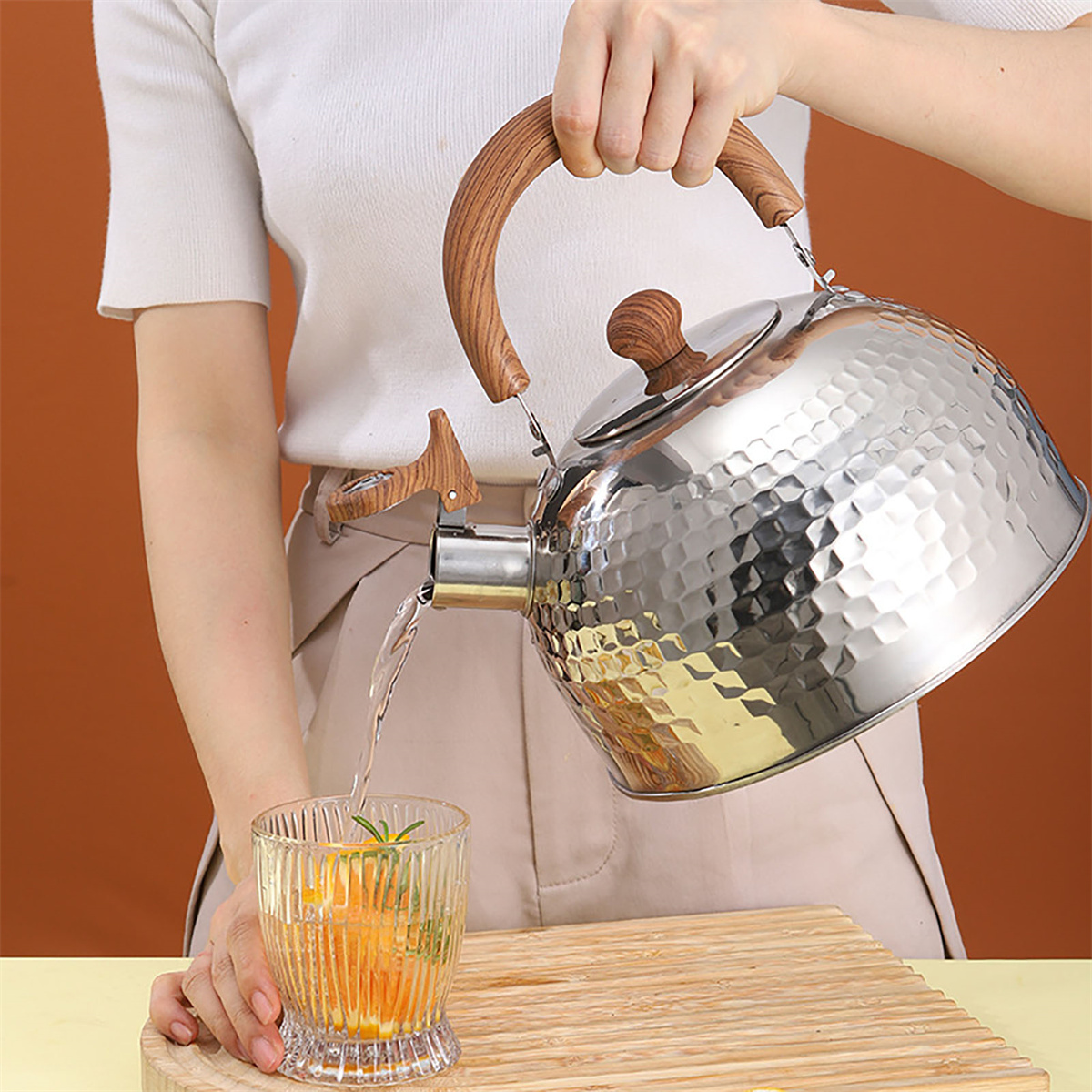Eine Person gießt Wasser aus einem silbernen Wasserkocher in ein Glas mit Orangenscheiben und Kräutern. Holzgriff und -sockel.