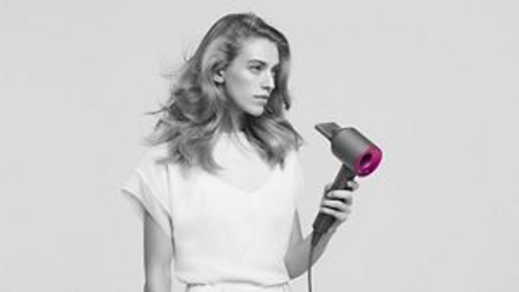 Eine Frau mit welligem Haar hält einen grauen und rosa Haartrockner. Sie trägt ein weißes Oberteil und blickt leicht nach links.