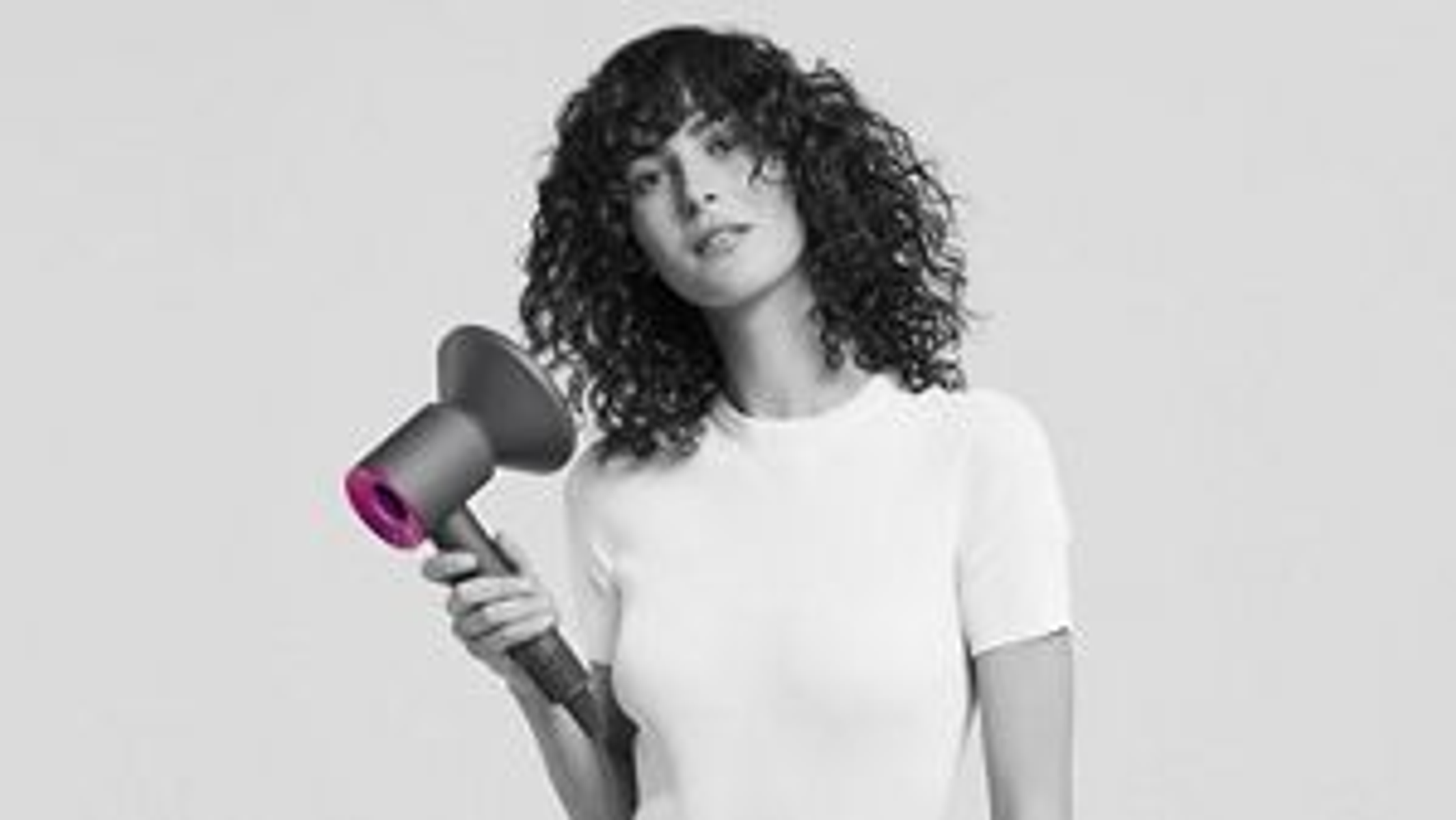 Eine Frau mit lockigem Haar hält einen grau-rosa Haartrockner. Sie trägt ein weißes Hemd vor grauem Hintergrund.