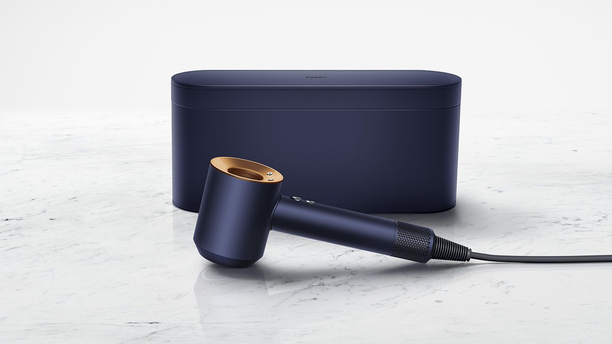 Ein marineblauer Dyson-Haartrockner mit goldfarbener Düse, dahinter eine Hülle, auf einer Marmoroberfläche. Schwarzes Kabel.