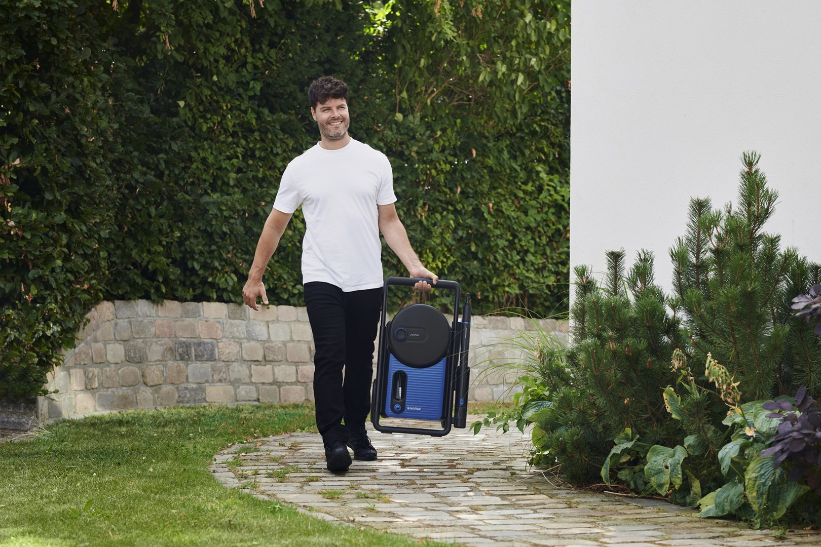 Mężczyzna w białej koszuli i czarnych spodniach niesie niebiesko-czarną myjkę ciśnieniową. Idzie po kamiennej ścieżce.