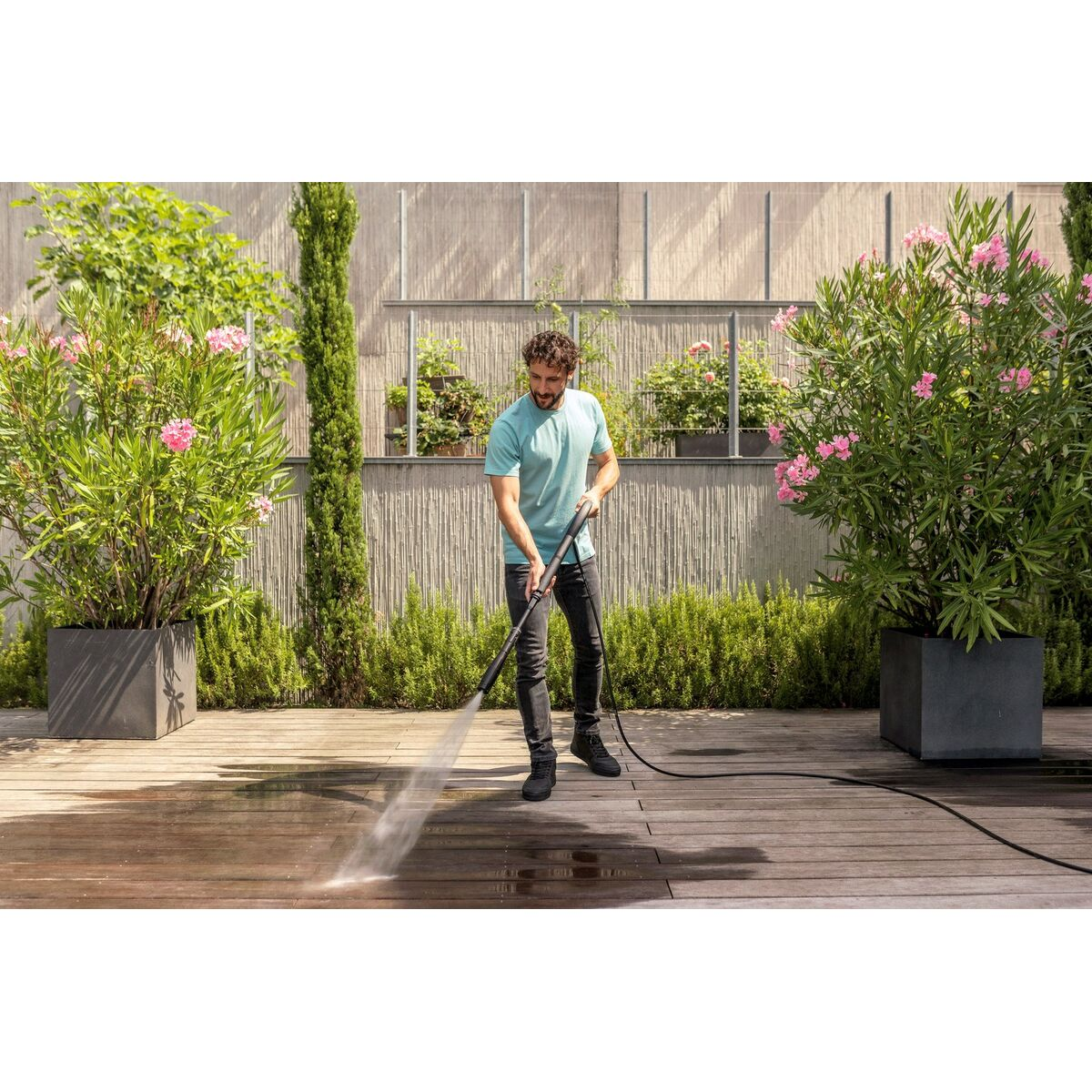Mężczyzna w niebieskiej koszuli myje drewniany taras myjką ciśnieniową. Rośliny i kwiaty w donicach w tle.