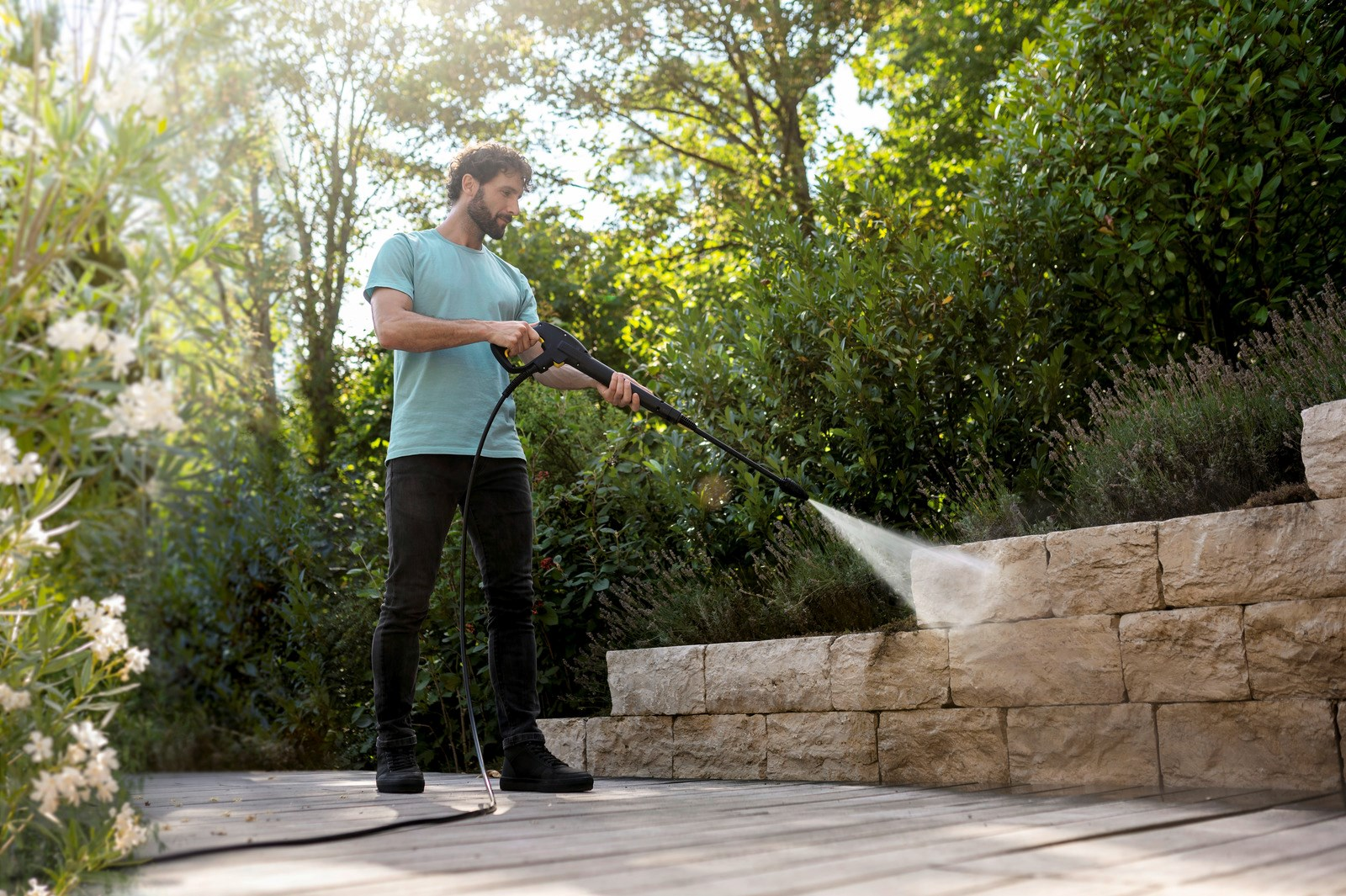 Mężczyzna w niebieskiej koszuli używa myjki ciśnieniowej na kamiennym murze w ogrodzie. Zielone rośliny i drzewa w tle.