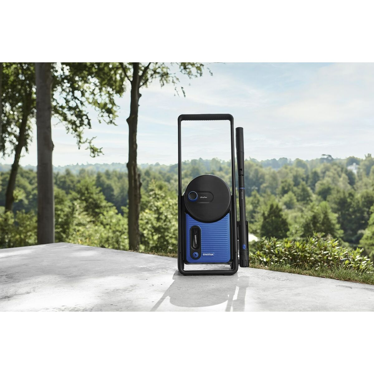 Niebiesko-czarna myjka ciśnieniowa stoi na betonowej powierzchni. Drzewa i zieleń wypełniają tło pod błękitnym niebem.