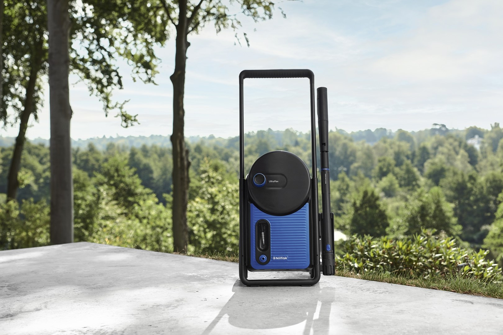 Niebiesko-czarna myjka ciśnieniowa stoi na patio z drzewami w tle. Niebo widoczne jest nad drzewami.
