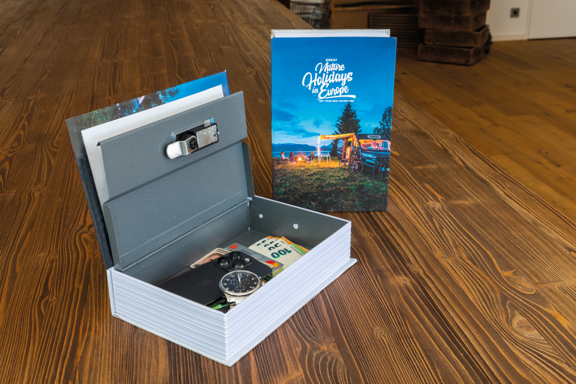 Eine graue Box mit Schloss ist geöffnet und zeigt Bargeld, eine Uhr und ein Telefon. Daneben liegt ein Buch mit einer Reiseszene auf einem Holztisch.