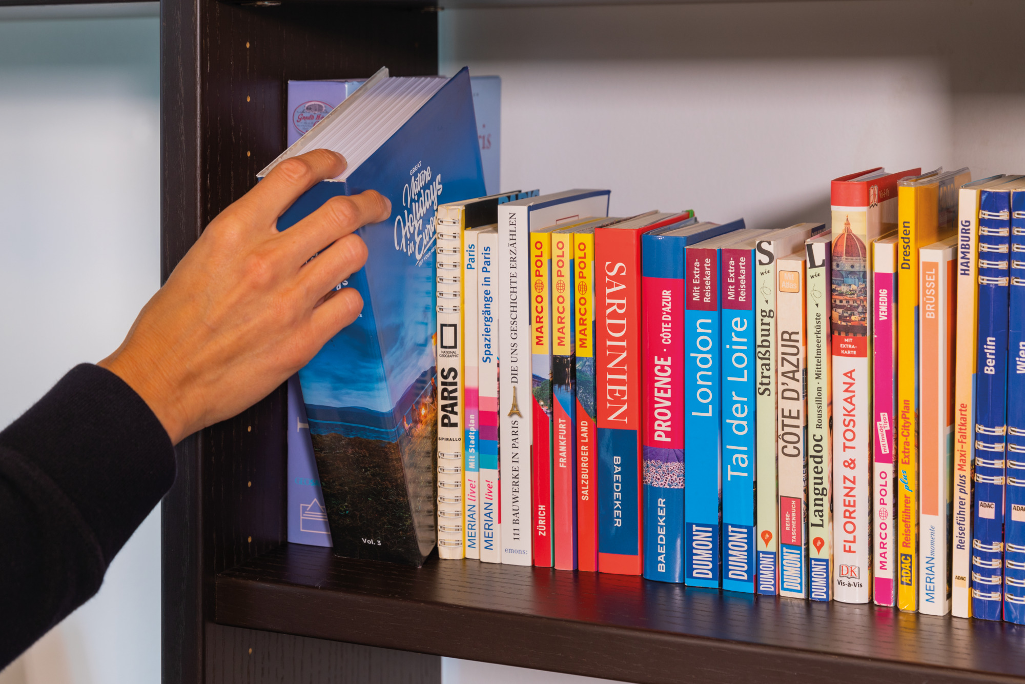 Eine Hand wählt ein blaues Buch aus einem Regal voller Reiseführer verschiedener Farben und Titel aus.