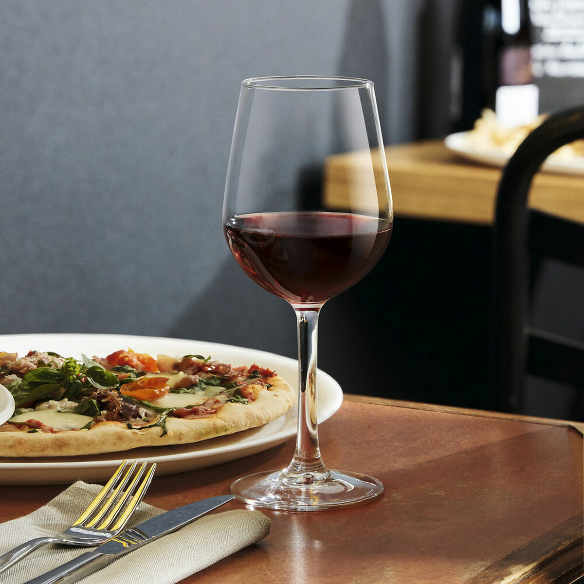 Ein Glas Rotwein steht vor einer Pizza auf einem weißen Teller. Gabel und Messer liegen auf einer Serviette. Der Tisch ist braun.