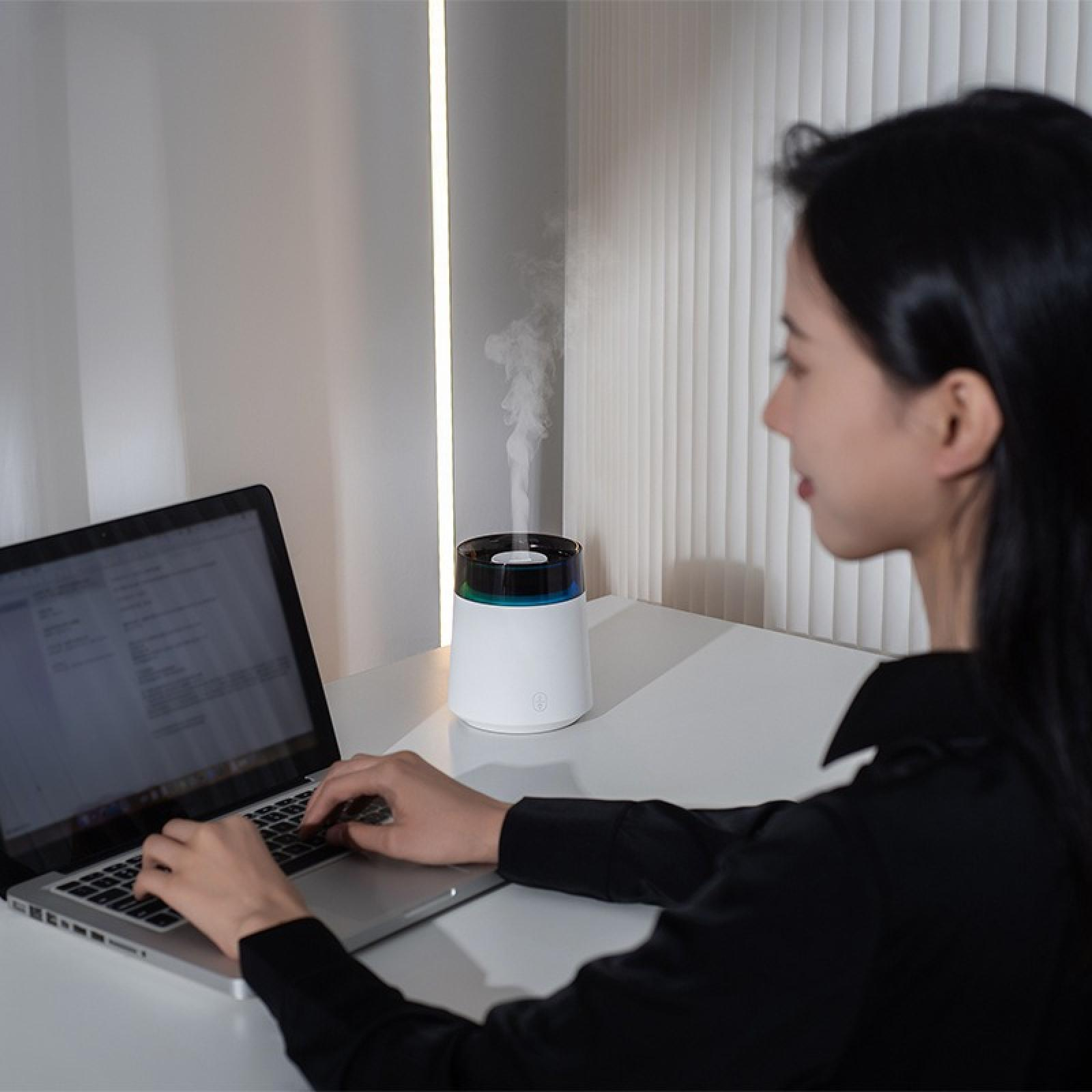 Eine Frau arbeitet an einem Laptop. Ein Luftbefeuchter steht auf dem Schreibtisch und gibt Dampf ab. Der Hintergrund hat Jalousien und eine Wand.