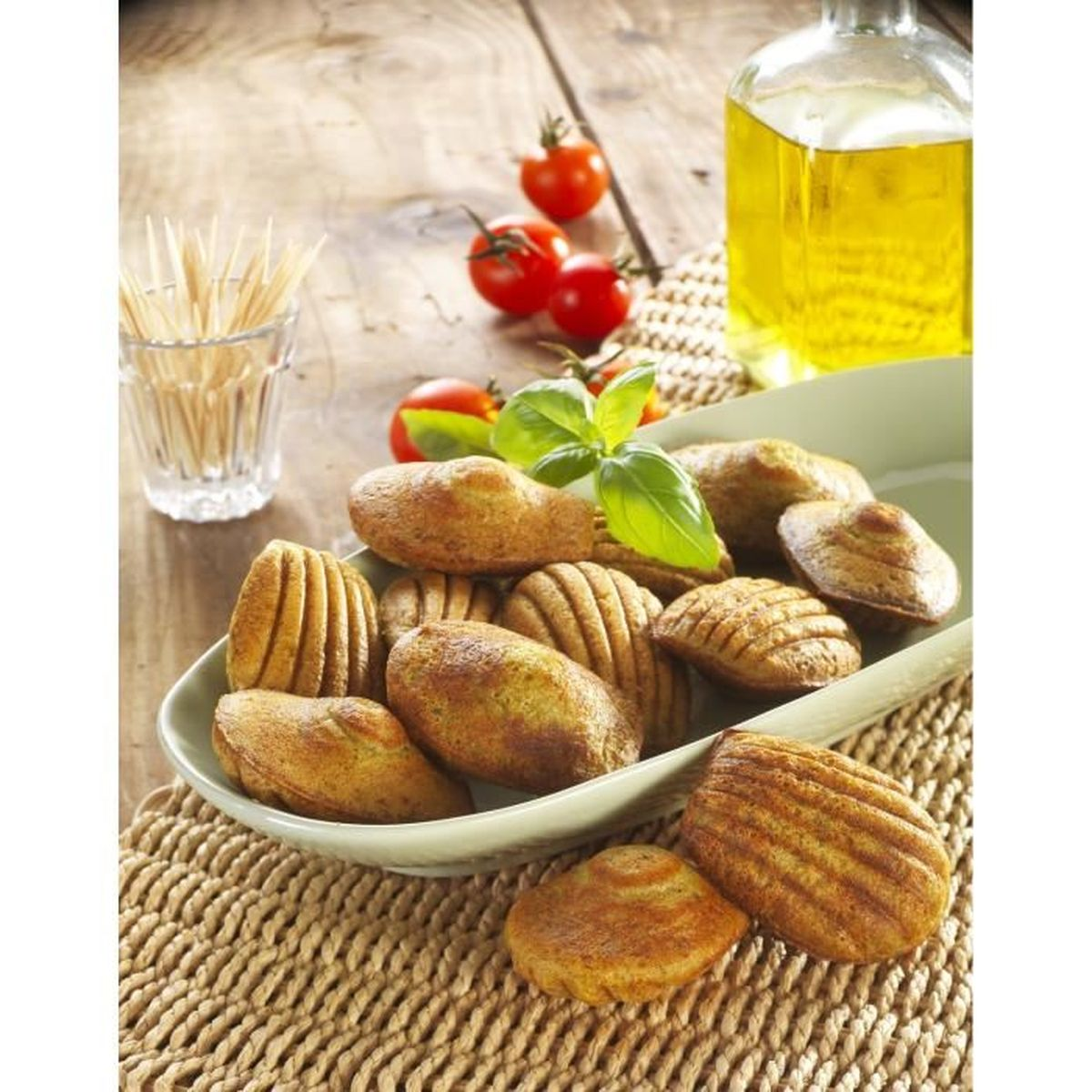 Madeleines in einer Schale, Basilikum, Tomaten, Ölflasche und Zahnstocher auf einem Holztisch mit gewebter Matte.