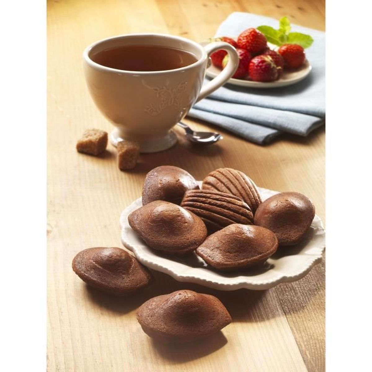 Eine Tasse Tee mit Madeleines auf einem Teller, Erdbeeren, Würfelzucker und einem Löffel, alles auf einem Holztisch.