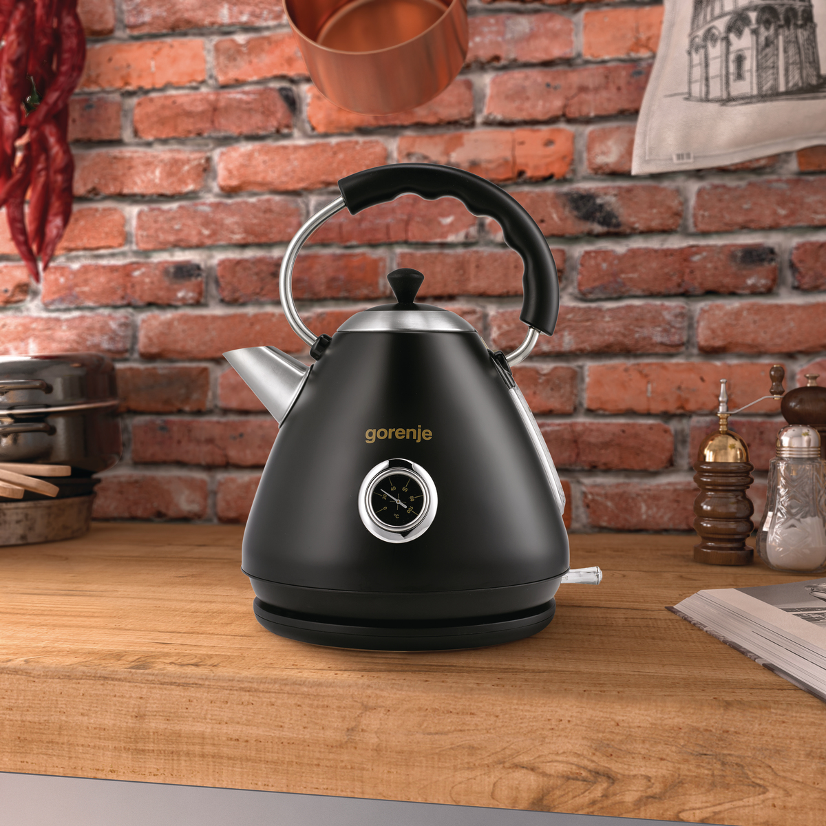 Un bollitore nero con indicatore di temperatura è su un piano di lavoro in legno. Un muro di mattoni fa da sfondo. Sono visibili utensili da cucina.