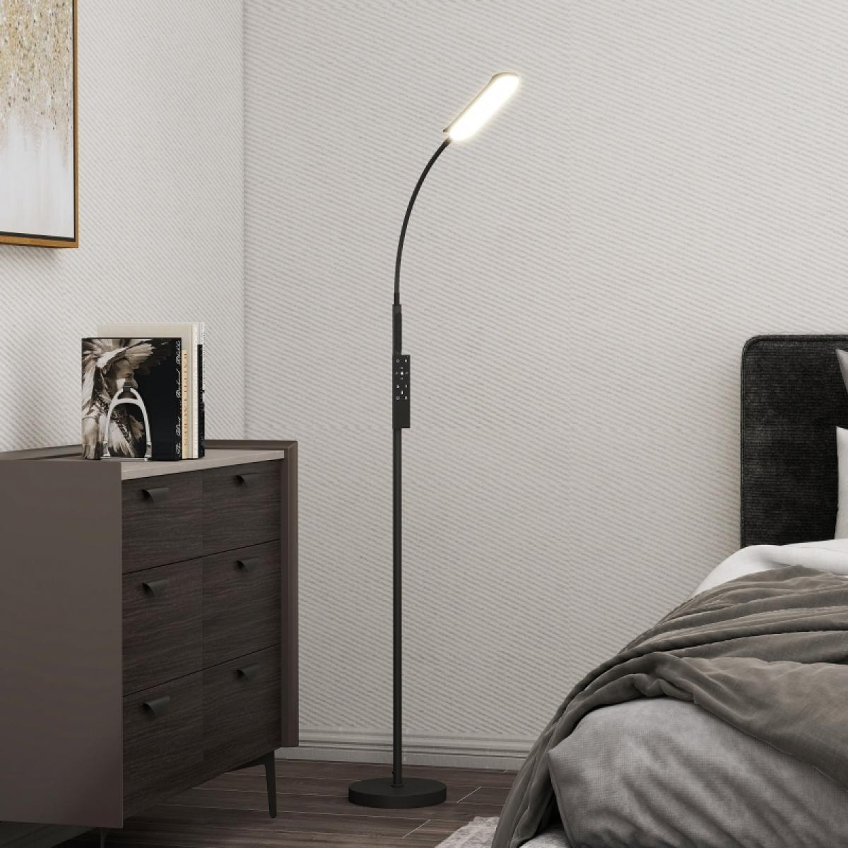 Eine schwarze Stehlampe leuchtet in einem Schlafzimmer. Eine Kommode mit Büchern steht neben der Lampe. Im Hintergrund ein Bett mit grauer Decke.