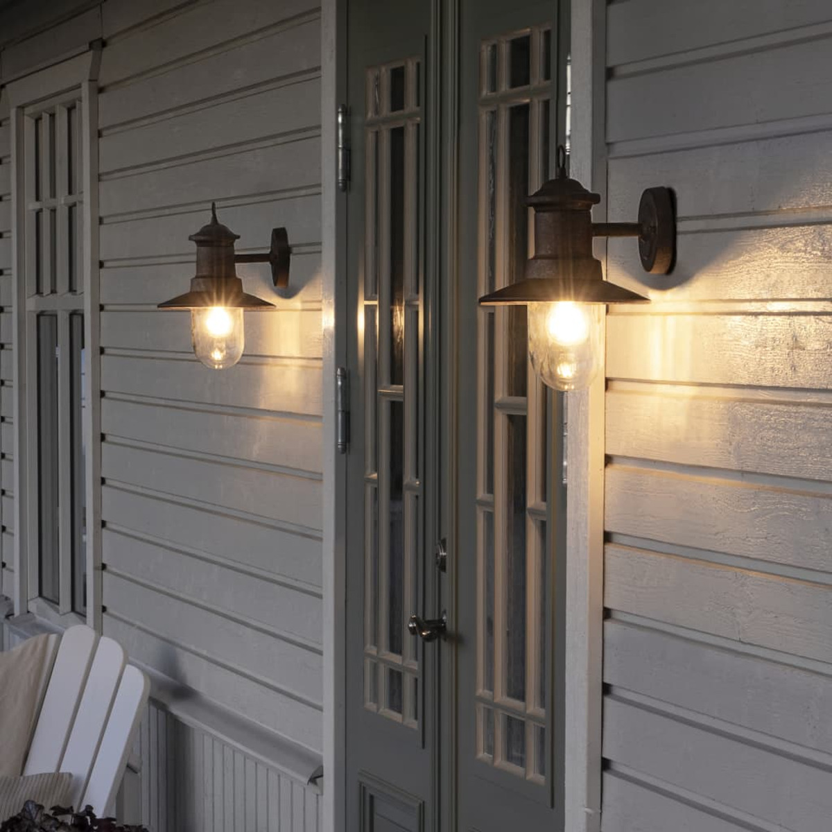Zwei Außenlampen beleuchten ein Holzhaus. Jede Lampe hat eine Glaskugel und einen dunklen Metallschirm. Eine Tür und ein Fenster sind sichtbar.