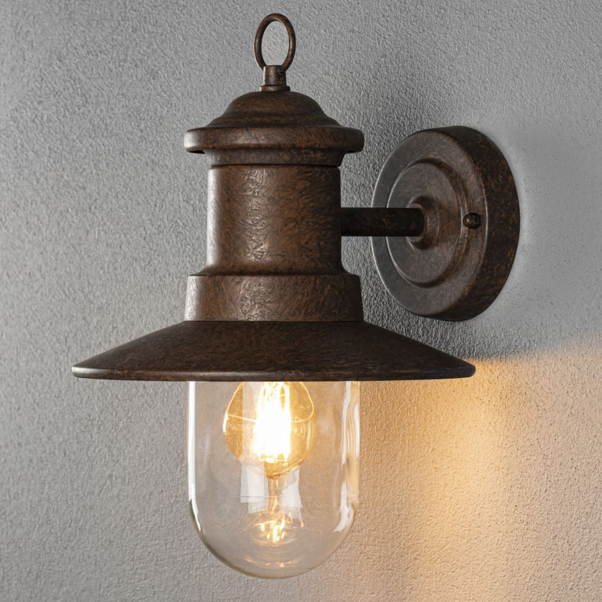Eine bronze-farbene Wandlampe mit Glühbirnenabdeckung, an einer strukturierten grauen Wand montiert. Die Glühbirne ist beleuchtet, die Lampe hat einen Ring.