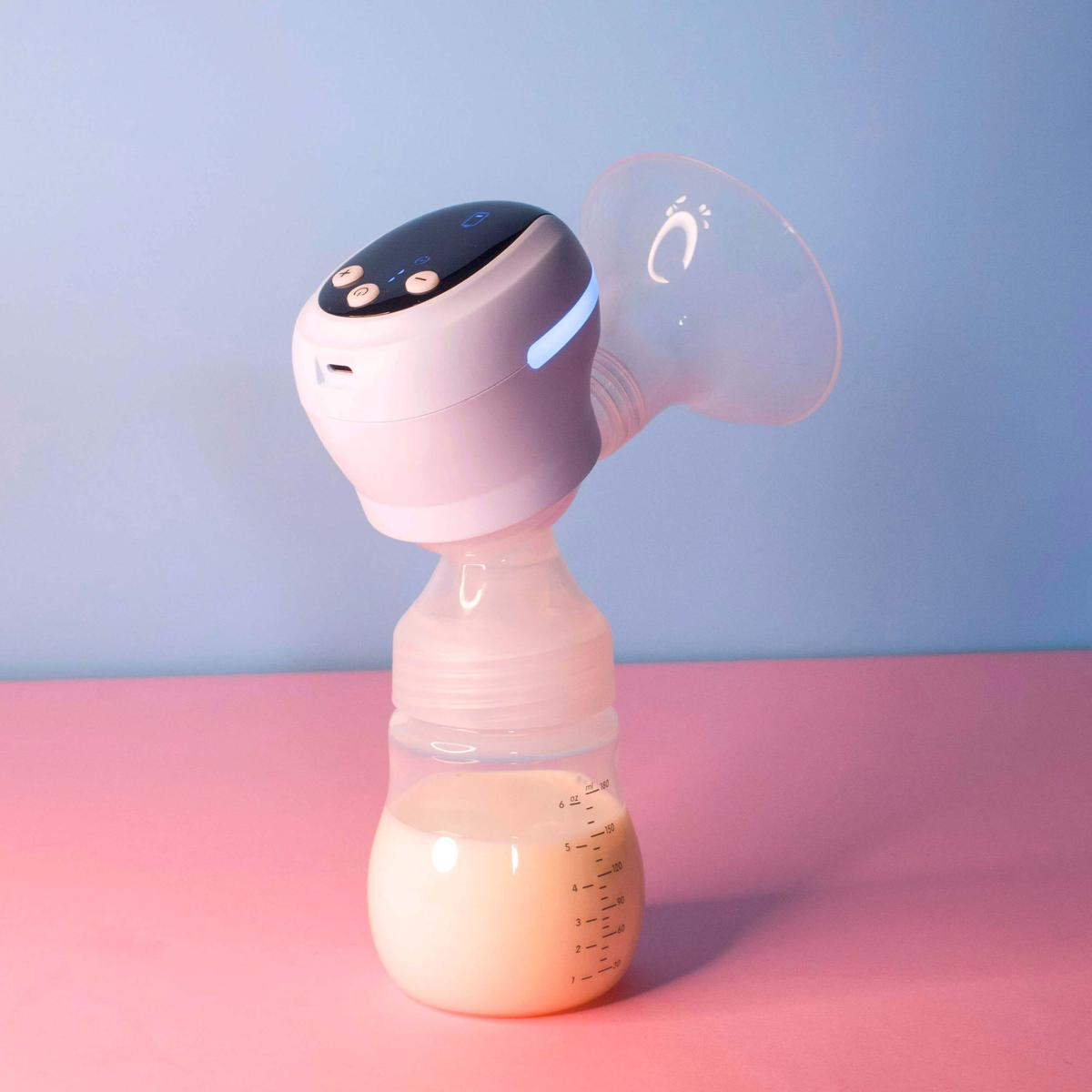 Laktator, składający się z białej maszyny i butelki, stoi na różowej powierzchni na tle niebieskiego. Mleko jest widoczne w butelce.