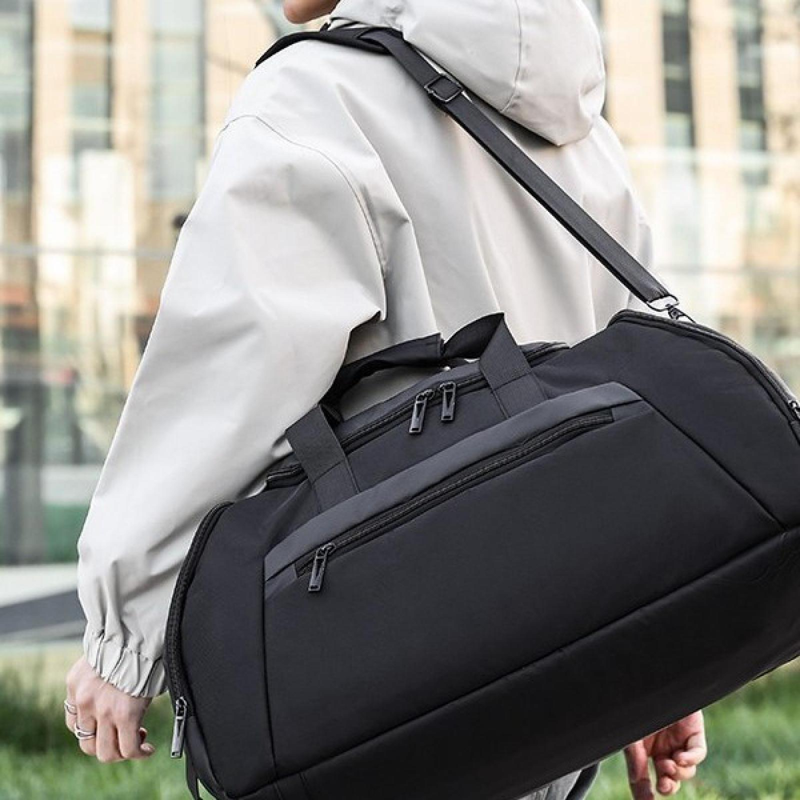 Eine Person in einer leichten Jacke trägt eine schwarze Reisetasche über der Schulter. Der Hintergrund zeigt ein Gebäude.