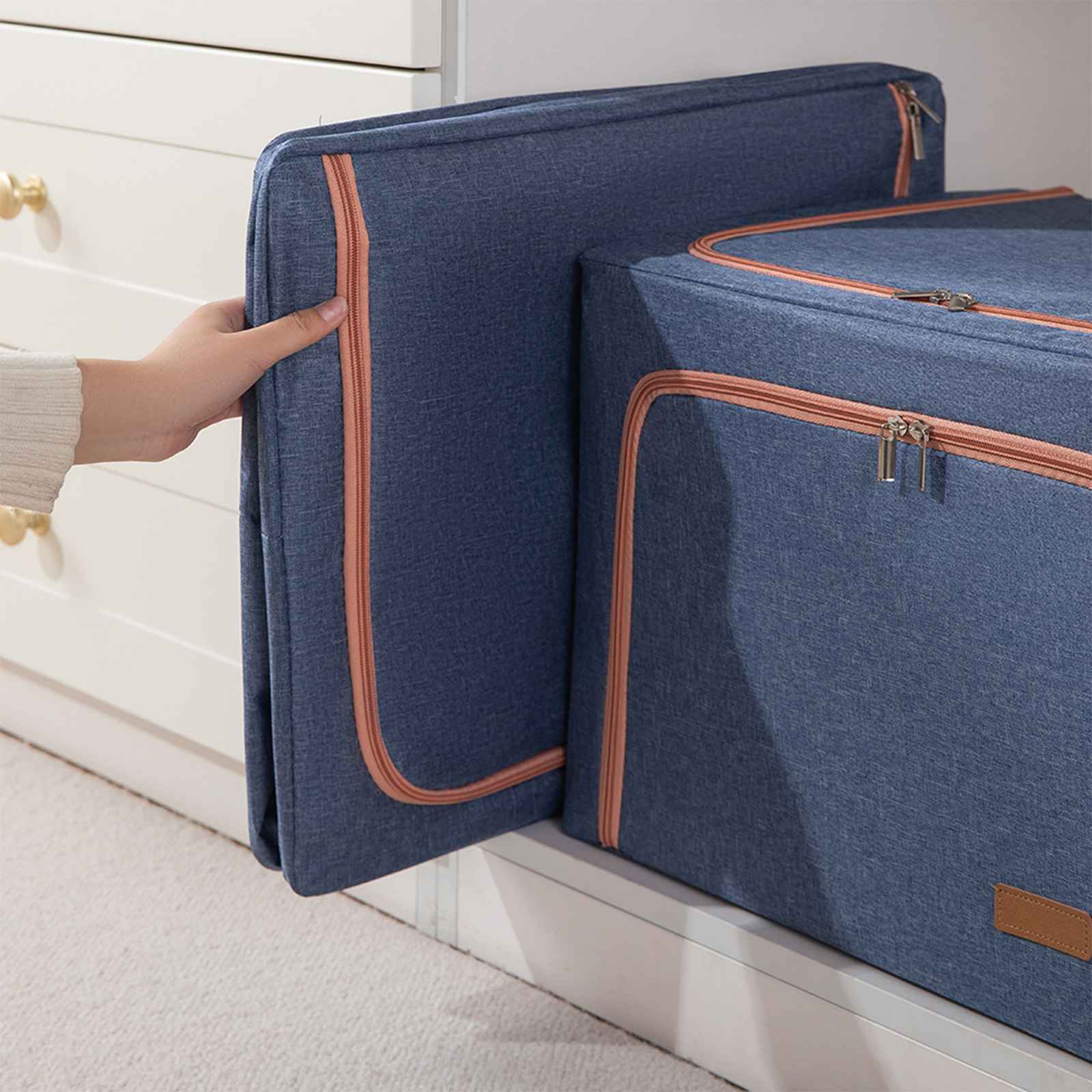 Eine Hand hält eine blaue Aufbewahrungsbox mit einem pfirsichfarbenen Reißverschluss. Eine andere Box ist dahinter. Beide stehen in einem weißen Schrank.