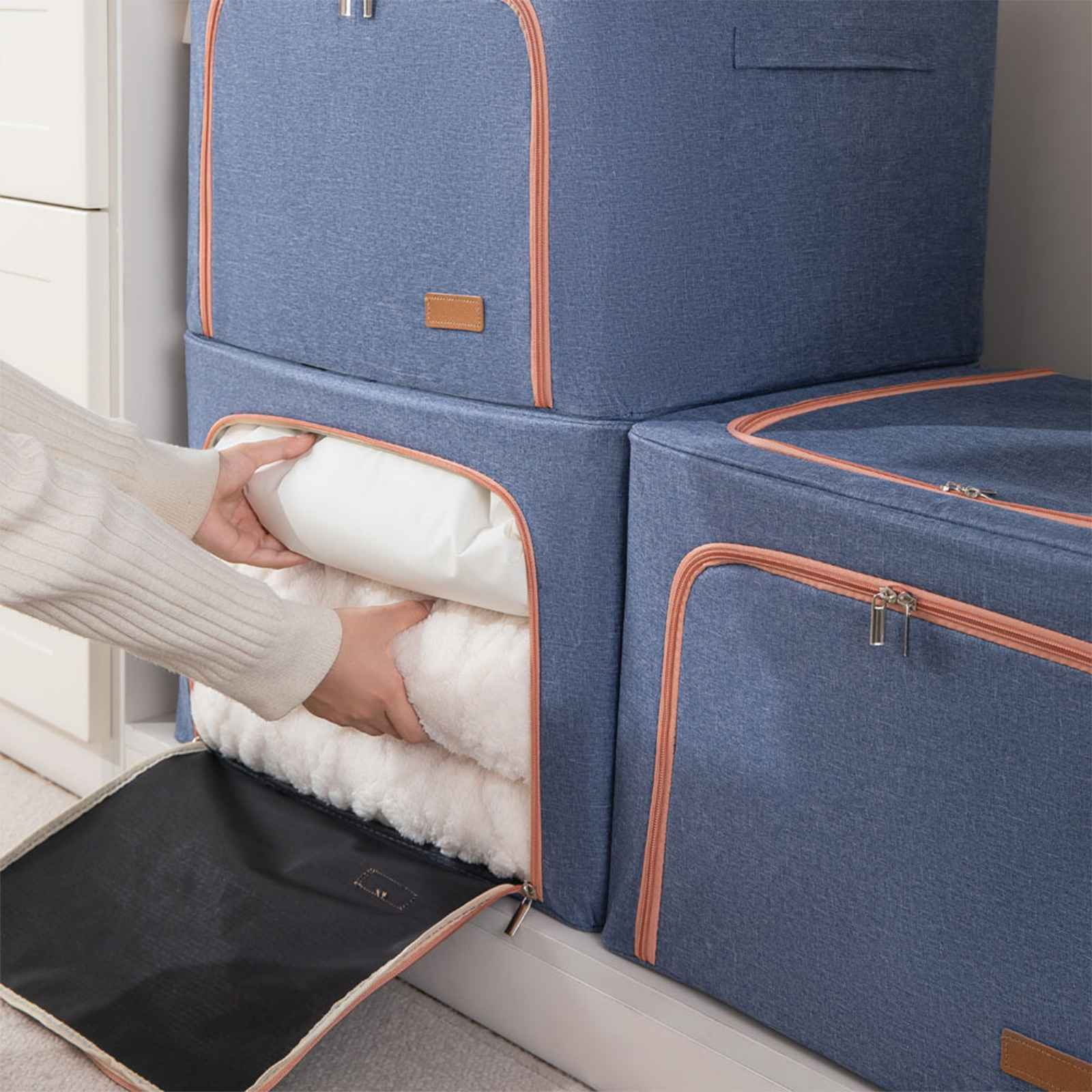 Eine Person öffnet eine blaue Aufbewahrungsbox und zeigt weiße Decken. Daneben sind weitere Boxen gestapelt.