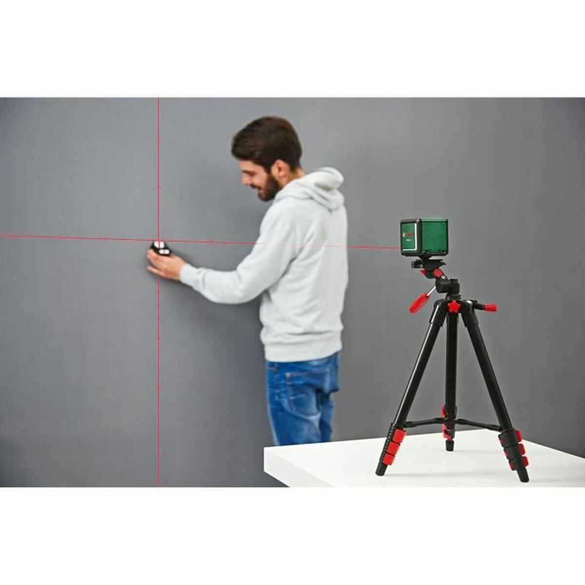 Bir adam duvarda lazer seviyesi kullanıyor. Bir tripod ve lazer masanın üzerinde. Kırmızı lazer çizgileri görülüyor.