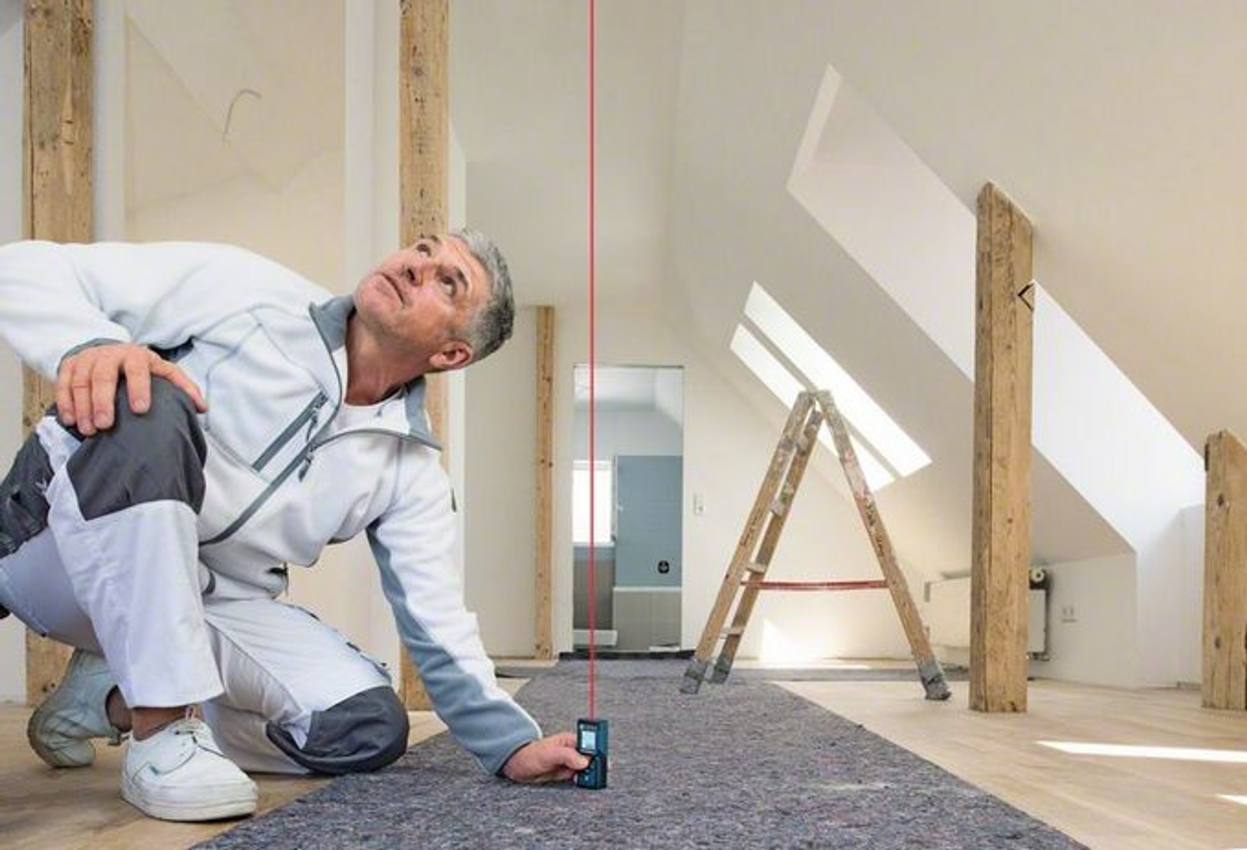 Mann kniet und benutzt einen Laser-Nivellierer. Eine Leiter und Holzbalken sind im Hintergrund. Die Szene spielt sich drinnen ab, wahrscheinlich bei einer Renovierung.