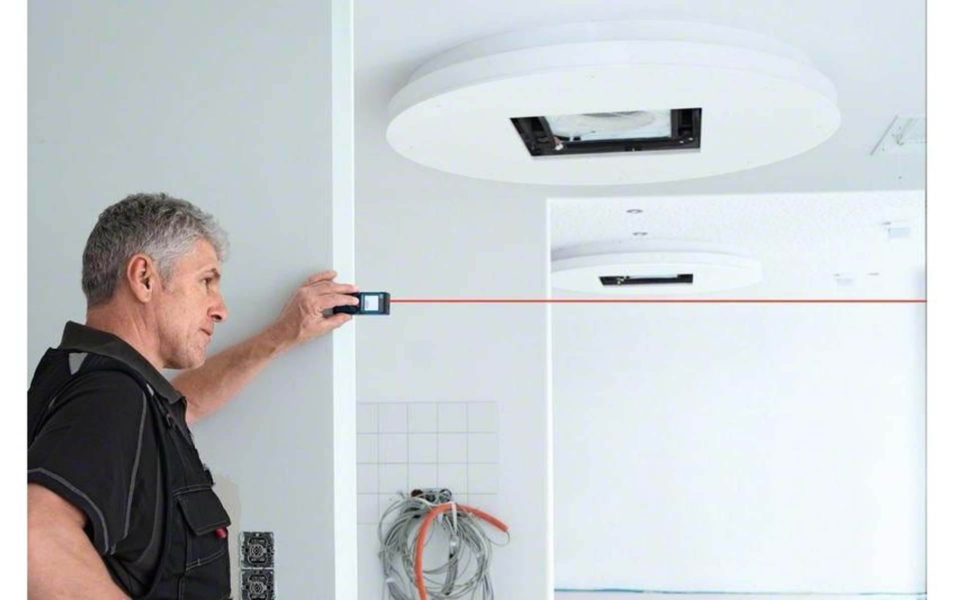 Ein Mann benutzt ein Laser-Messwerkzeug zum Messen der Entfernung. Es gibt eine weiße Decke mit einem eingelassenen Licht und einige elektrische Drähte.
