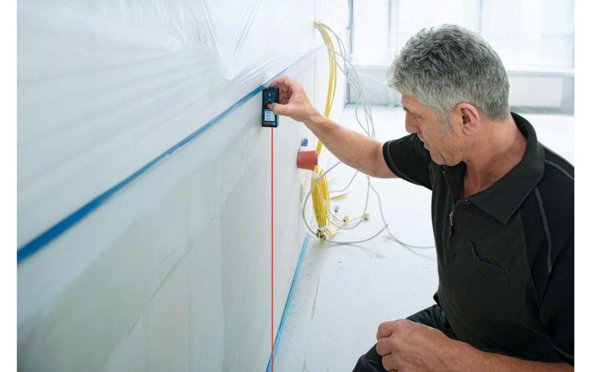 Ein Mann benutzt ein Laser-Entfernungsmessgerät an einer weißen Wand. Eine rote Laserlinie ist sichtbar. Drähte und ein Fenster sind im Hintergrund.