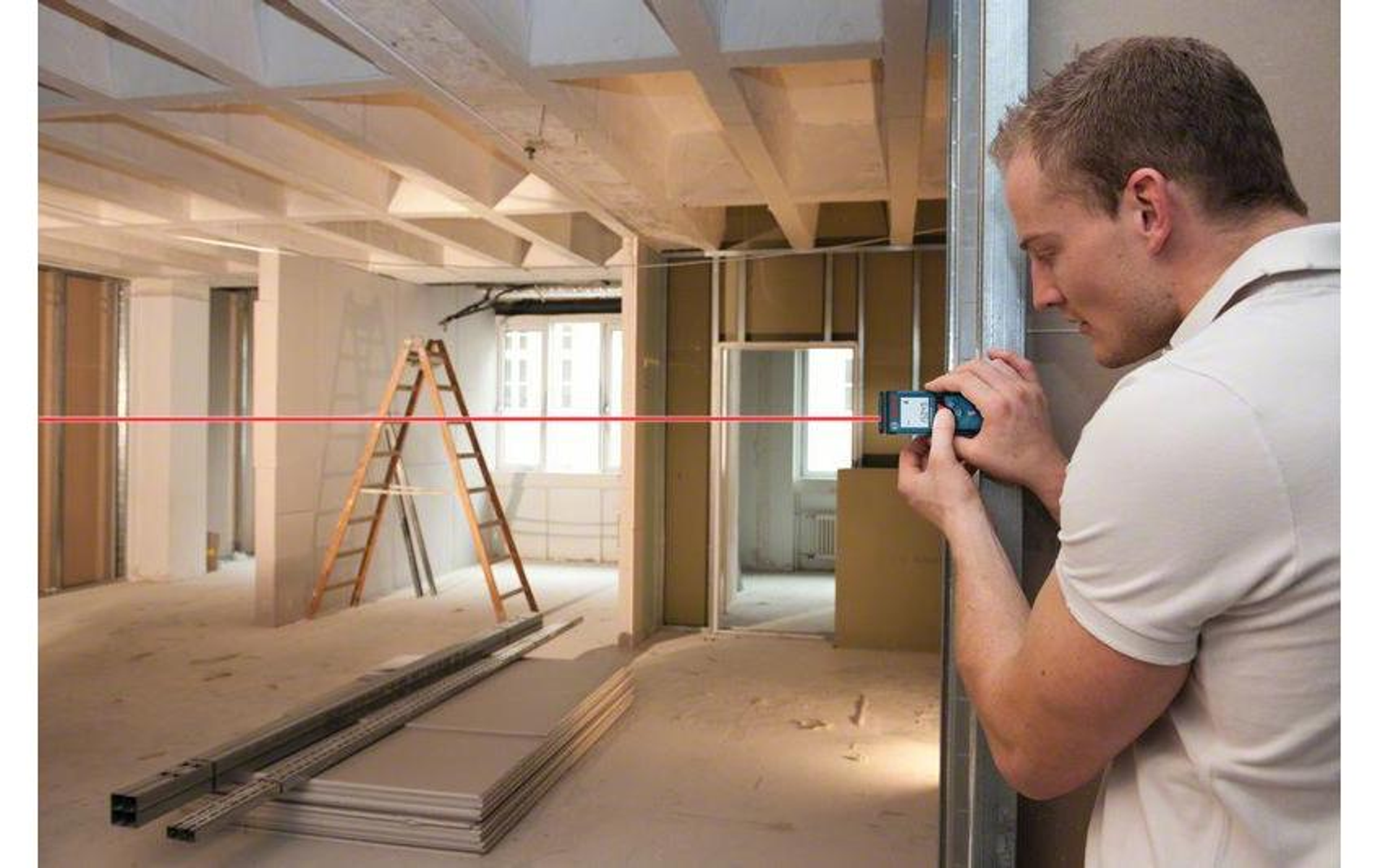 Ein Mann benutzt einen Laser in einer Baustelle. Ein roter Laserstrahl erstreckt sich über den Raum. Es gibt eine Leiter und Baumaterialien.