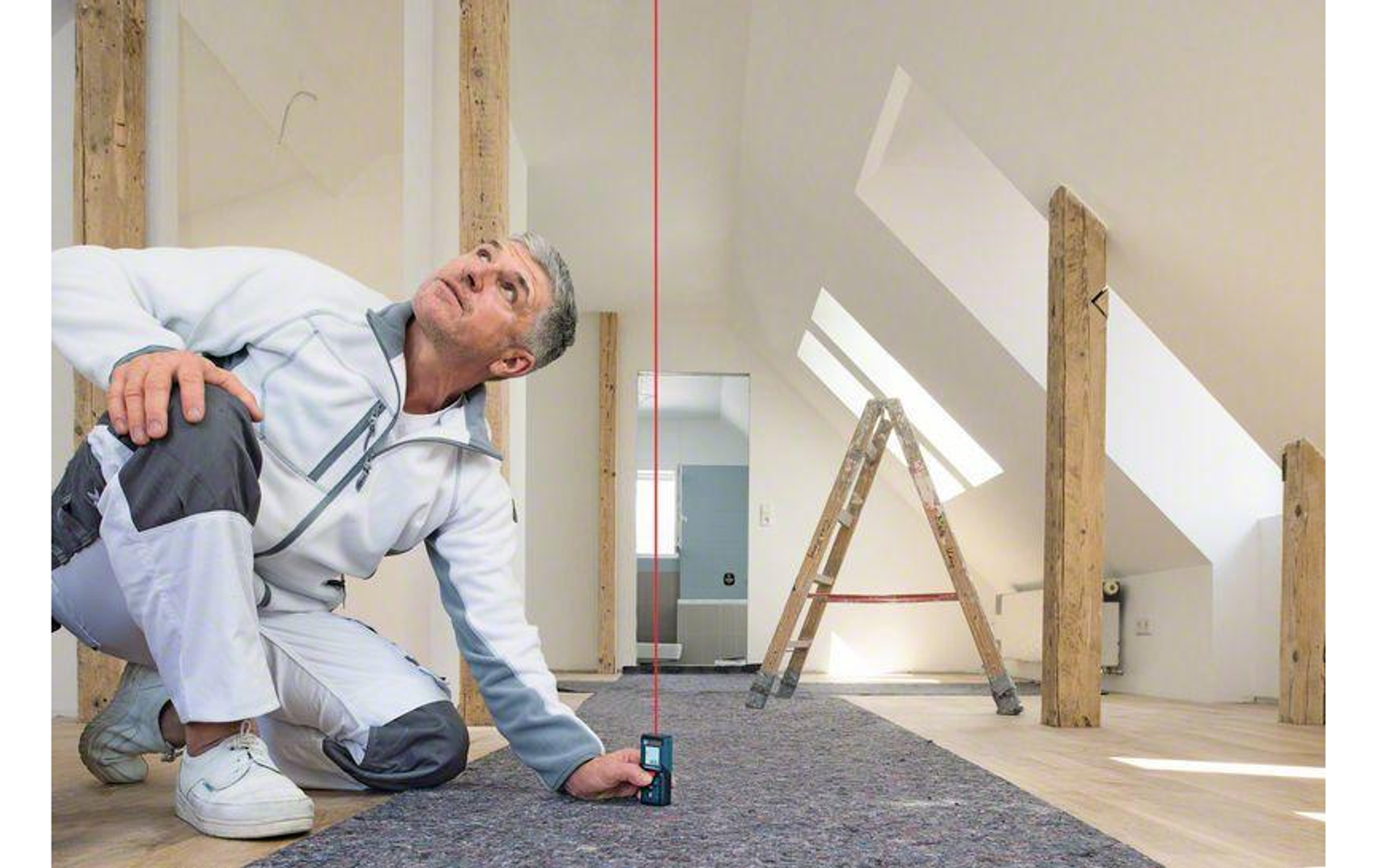 Ein Mann kniet und benutzt in einem Dachboden einen Laser. Roter Laserstrahl erstreckt sich nach oben. Leiter und Holzbalken sind sichtbar.