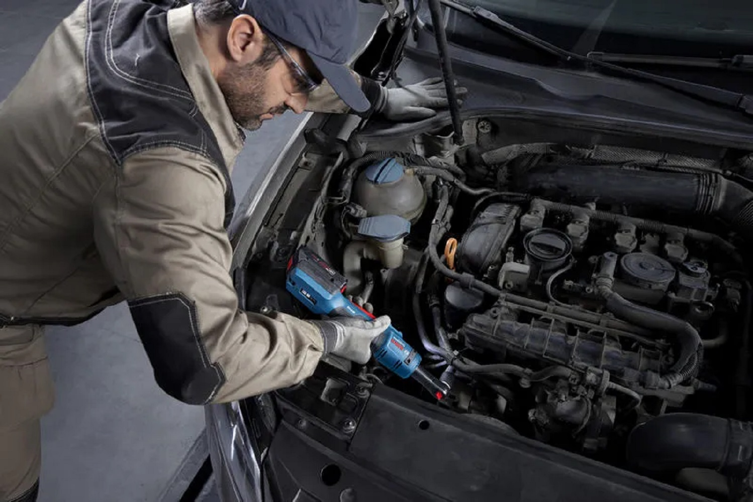 İş kıyafetleri ve şapka takmış bir tamirci, mavi ve siyah bir elektrikli el aleti kullanarak bir araba motorunu inceliyor. Motor bölmesi açık.